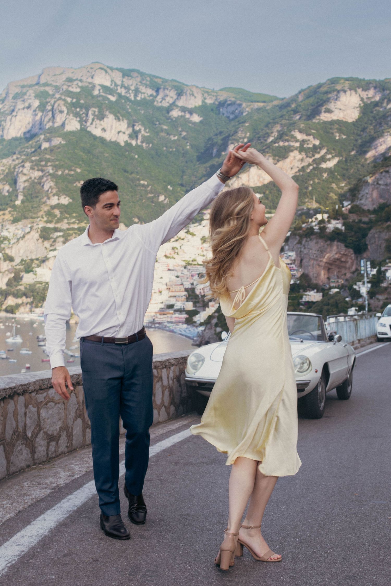 Couple dancing on Amalfi Coast road with woman in yellow dress spinning near vintage white convertible.