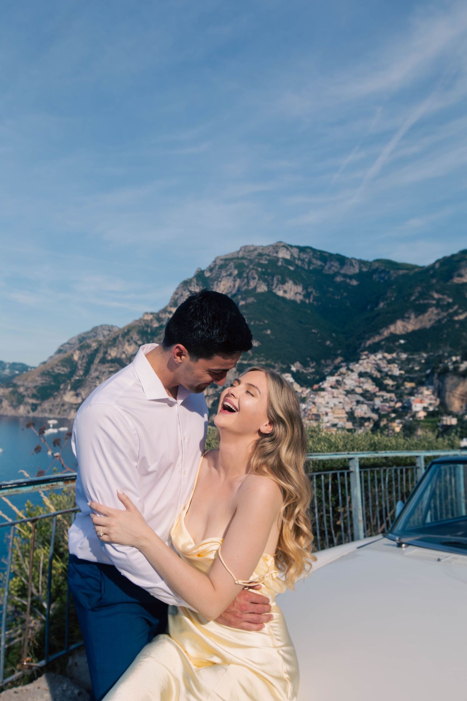 Joyful couple laughing and embracing by a white vintage car with Positano cliffs and blue sky behind them.