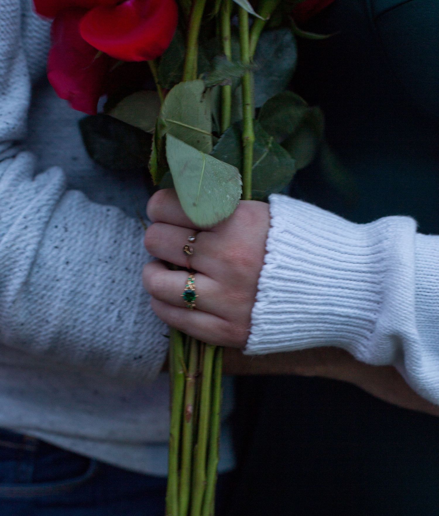 Hand holding red roses wearing a delicate gold ring with green emerald stone, white knit sweater sleeve visible.