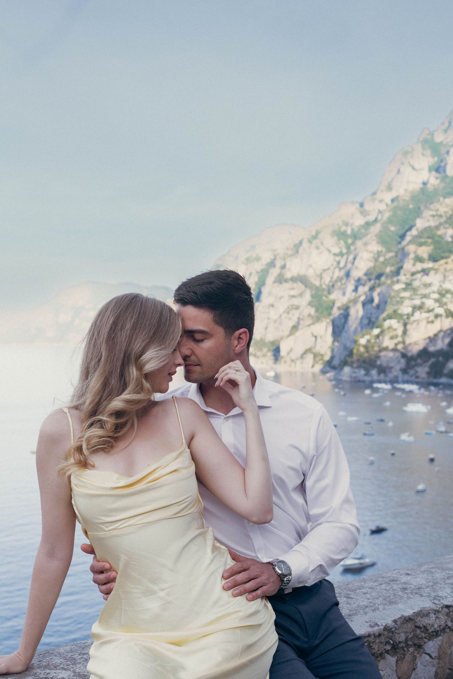 Romantic couple embracing on Amalfi Coast terrace, woman in yellow dress, man in white shirt, with sea and cliffs behind.