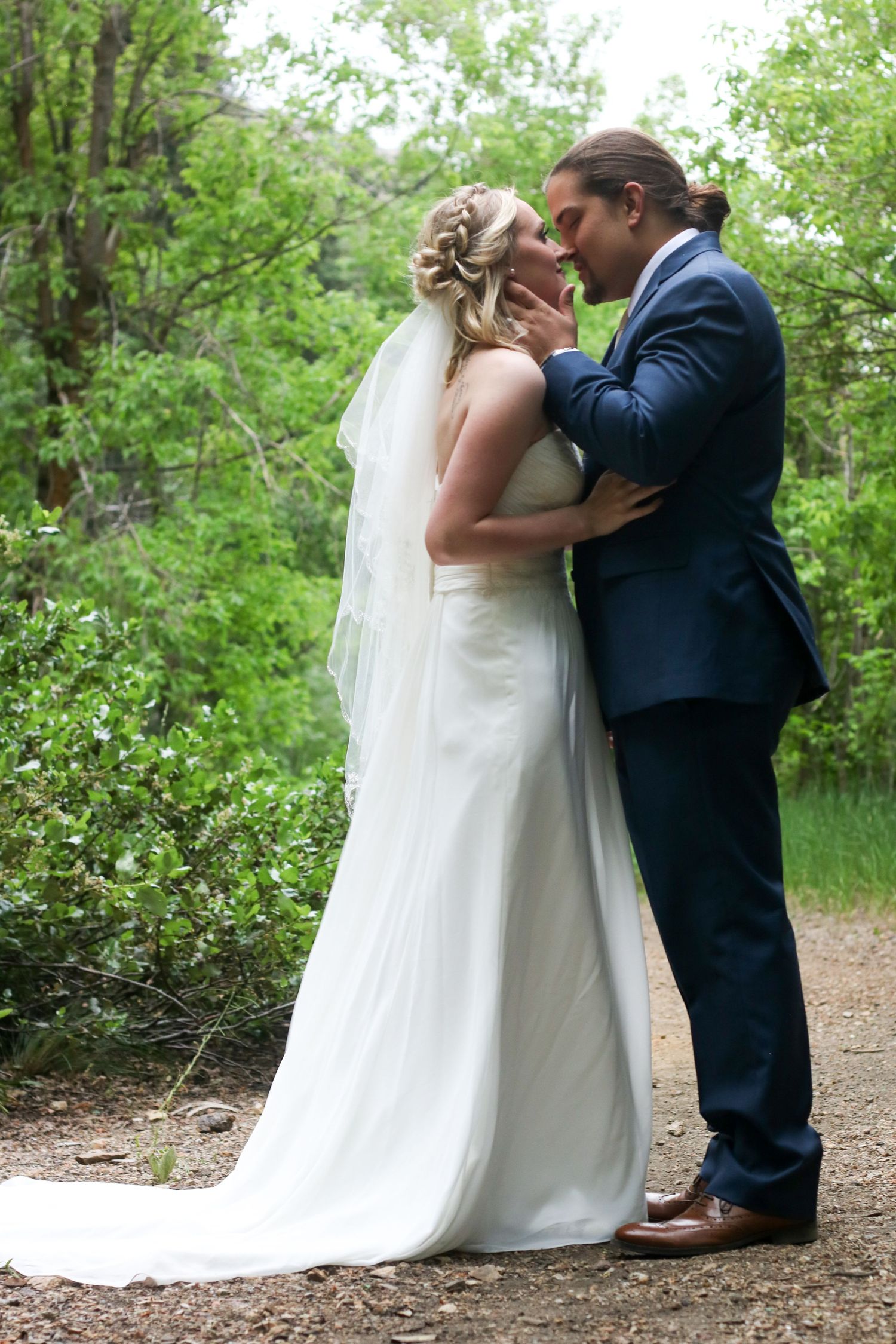 A tender wedding moment captured on a tree-lined nature path with flowing white dress and veil.