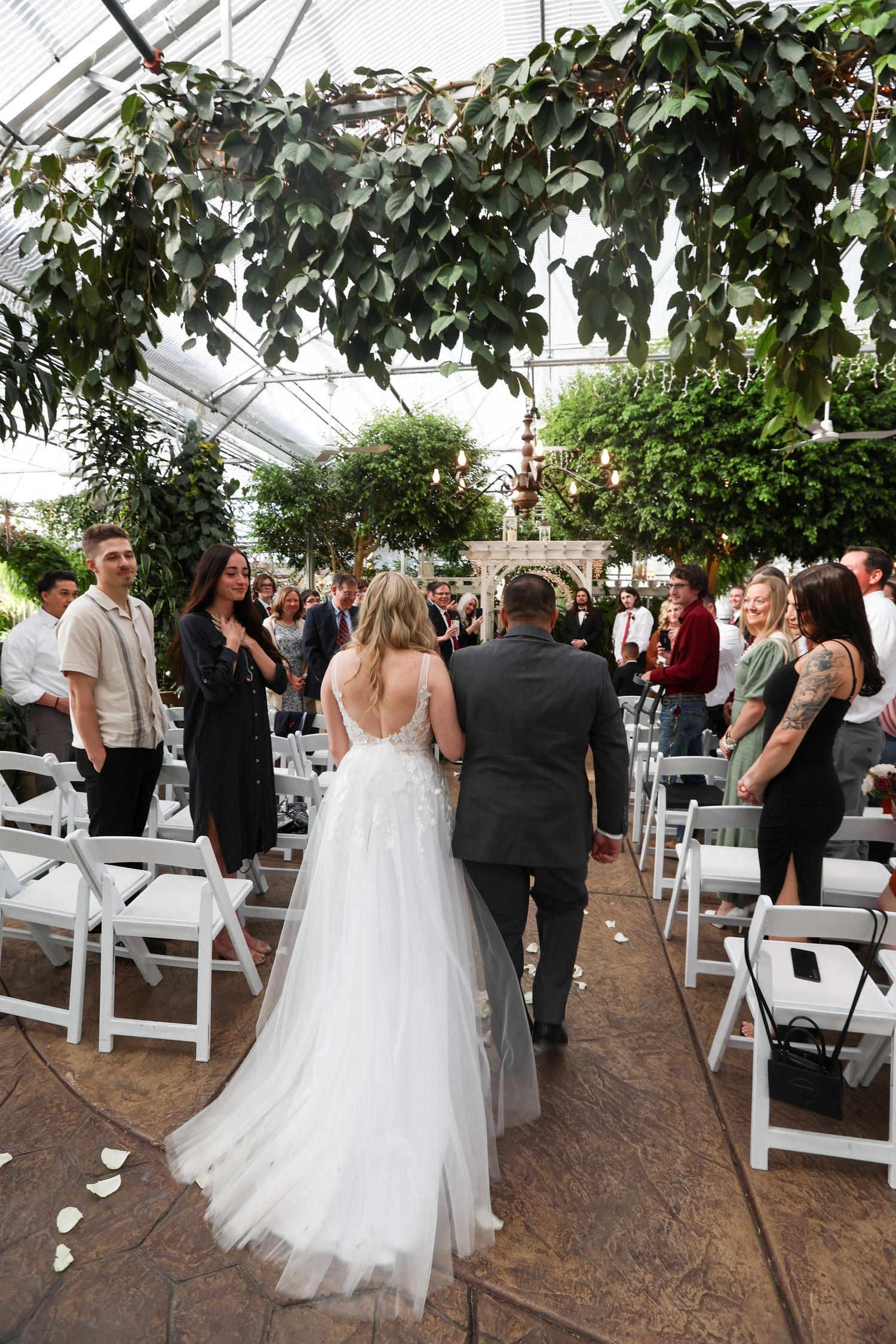 Wedding processional in a greenhouse venue with white chairs and hanging greenery.