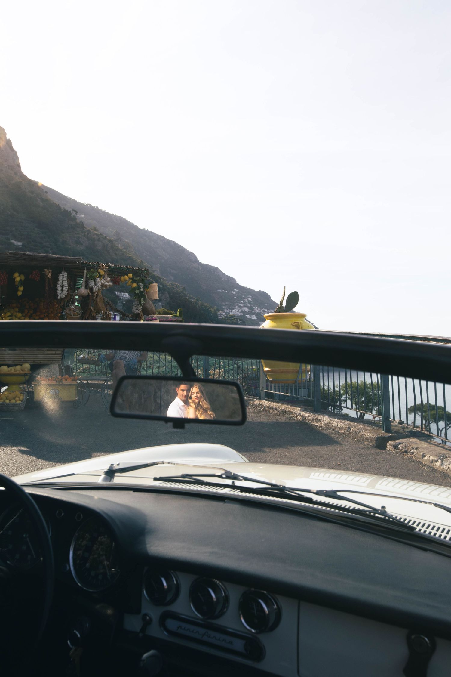 Couple reflected in vintage convertible car rearview mirror on scenic Amalfi Coast road in Italy.