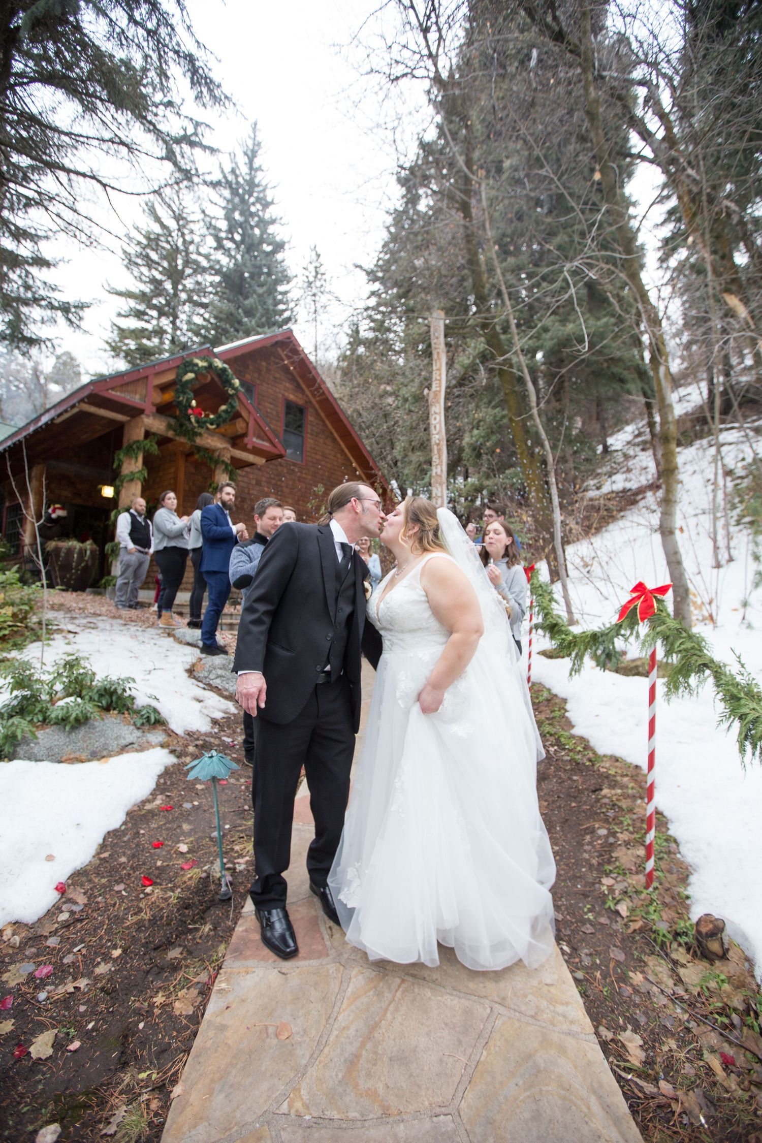 Bride and groom share first kiss on snowy winter wedding path outside rustic log cabin chapel.