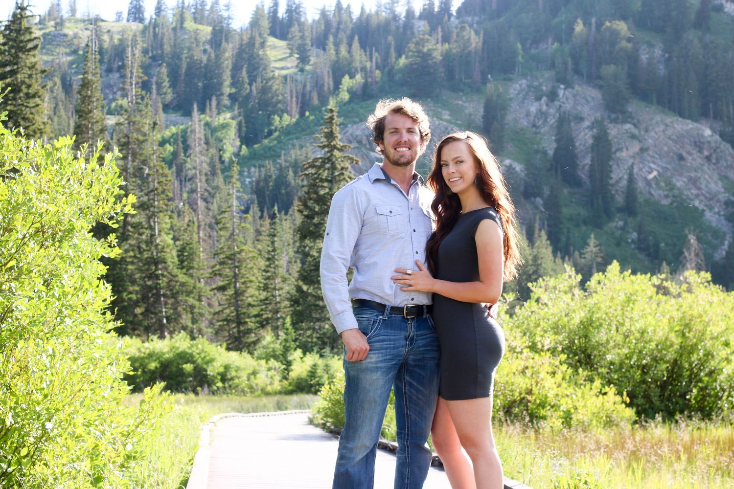 A couple poses together on a mountainside with evergreen forests and peaks in the background.