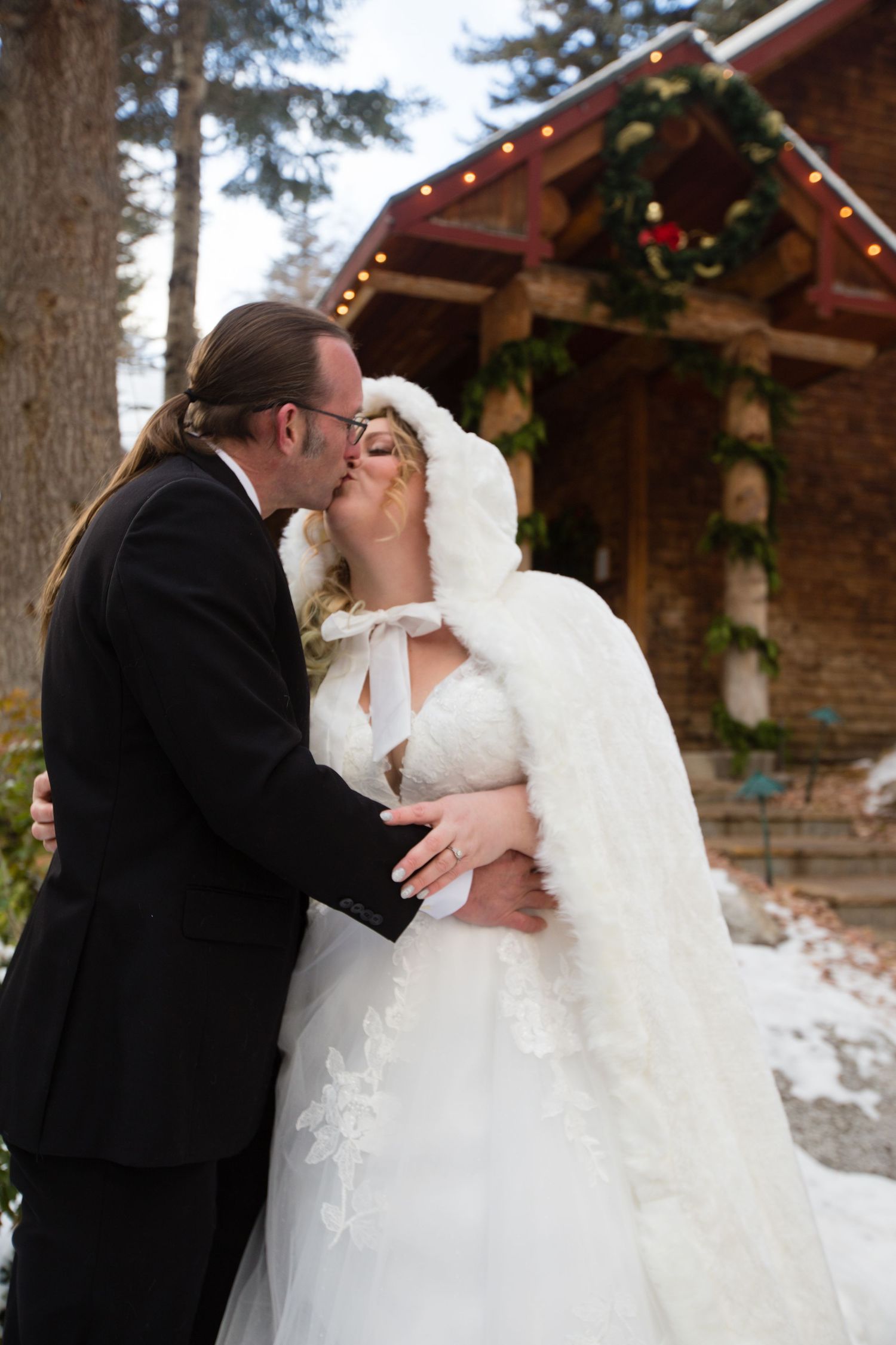 A romantic winter wedding kiss captured in front of a festively decorated cabin.