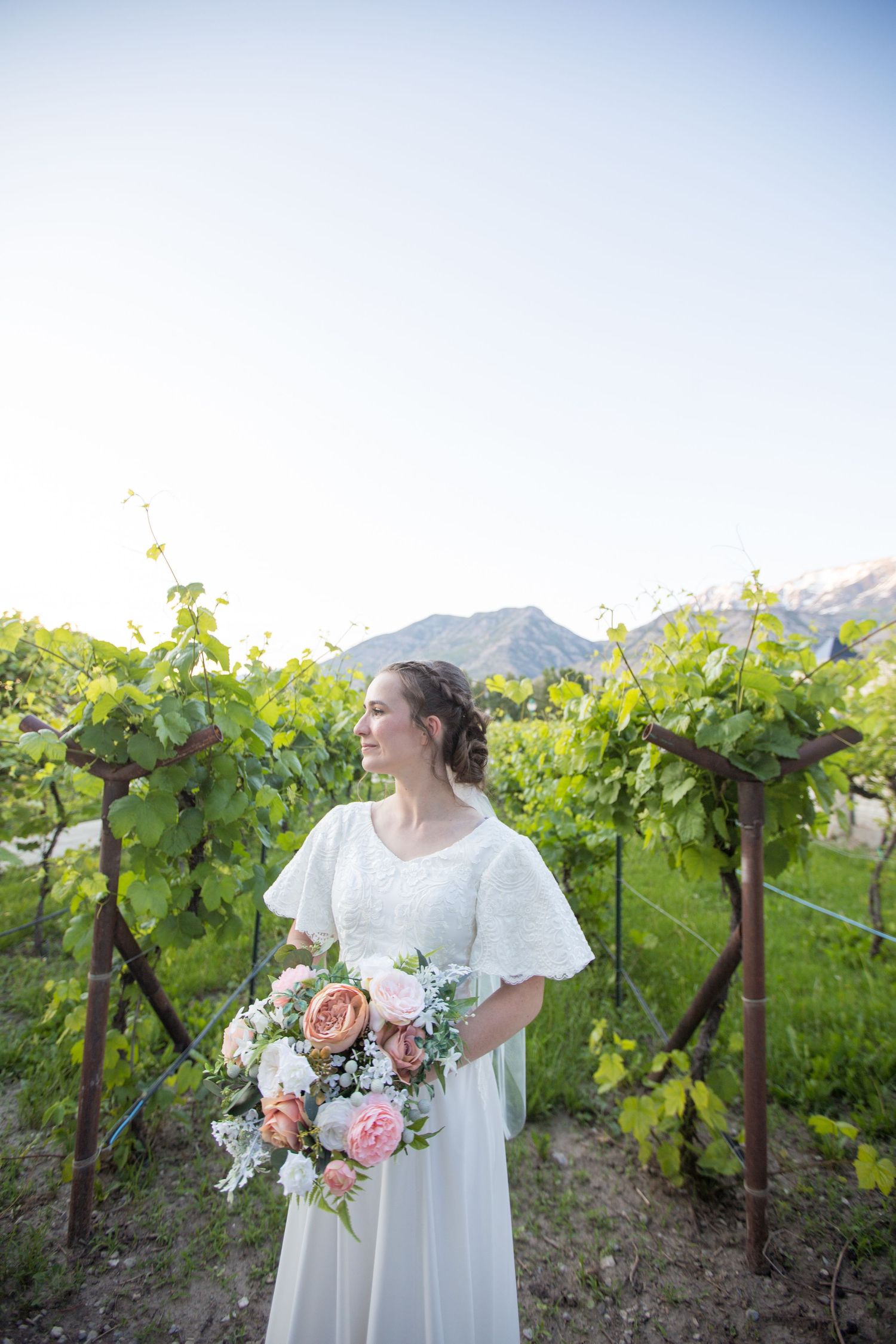 A white wedding dress and bouquet photographed in a scenic vineyard with mountains in the background.