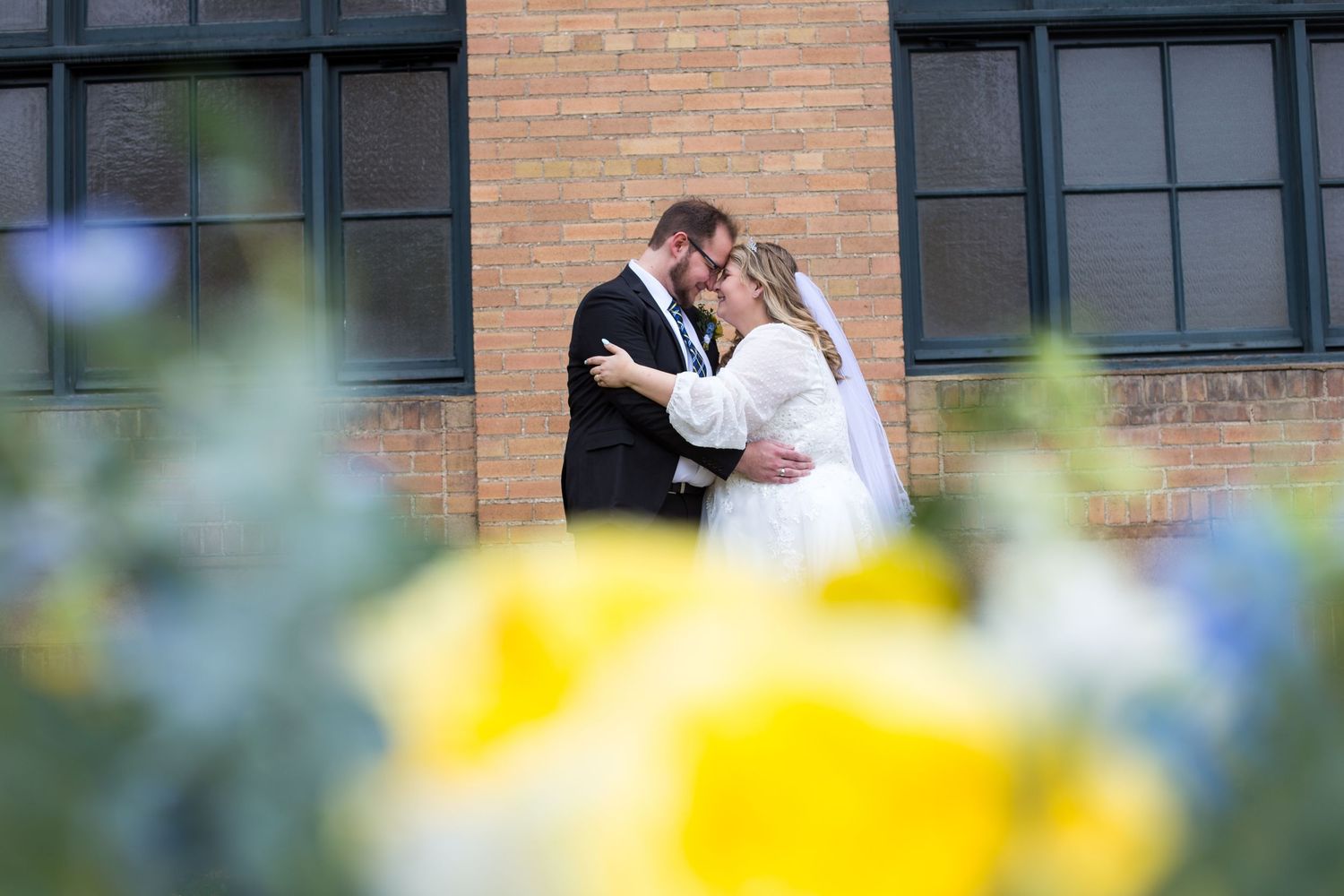 Bride and groom sharing a romantic kiss outdoors near brick building with yellow flowers in foreground.