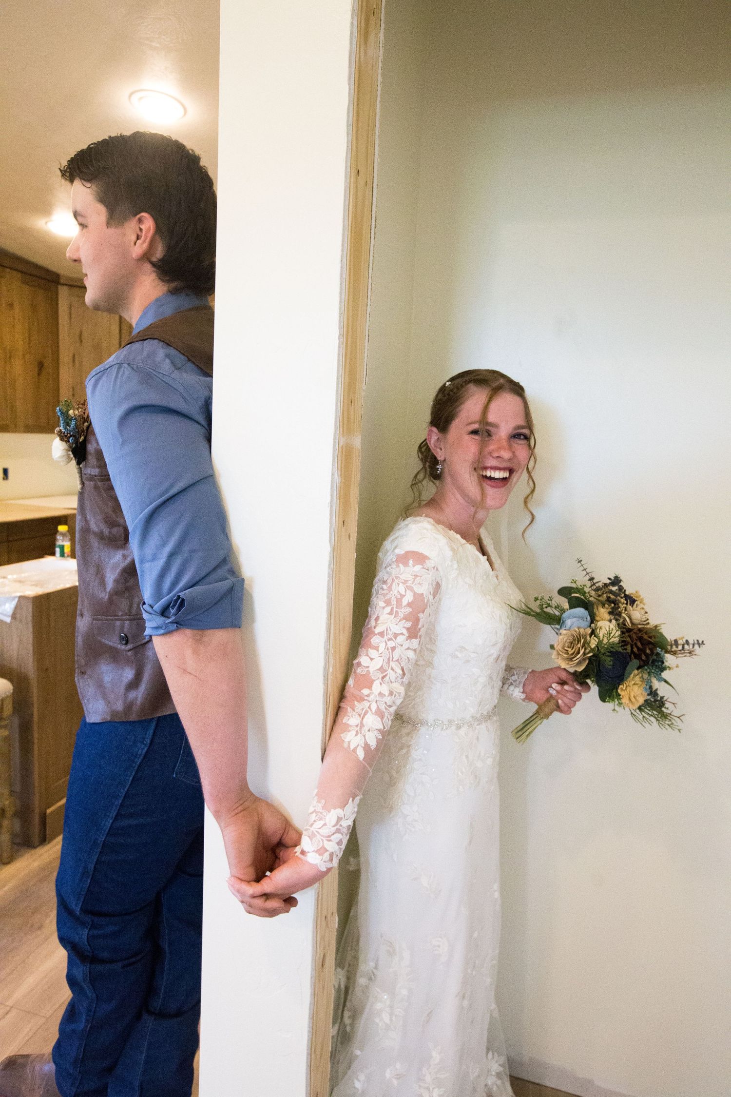 Bride in lace gown holds bouquet, laughing while holding hands with groom around a wall in a sweet first look moment.