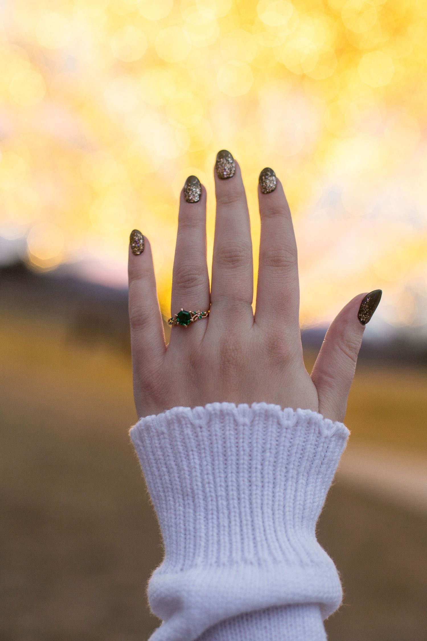 Hand with gold glitter nails and green gemstone ring raised against golden bokeh background.