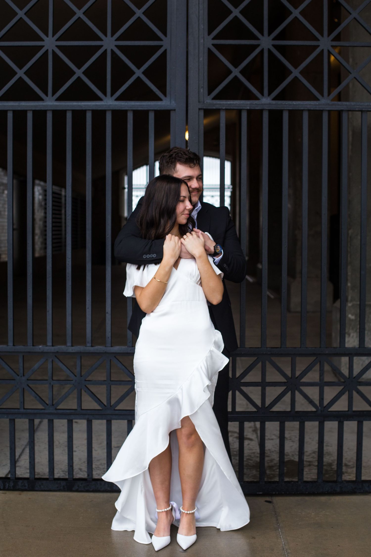 Bride in white ruffled gown embraced by groom in black suit in front of ornate iron gate.