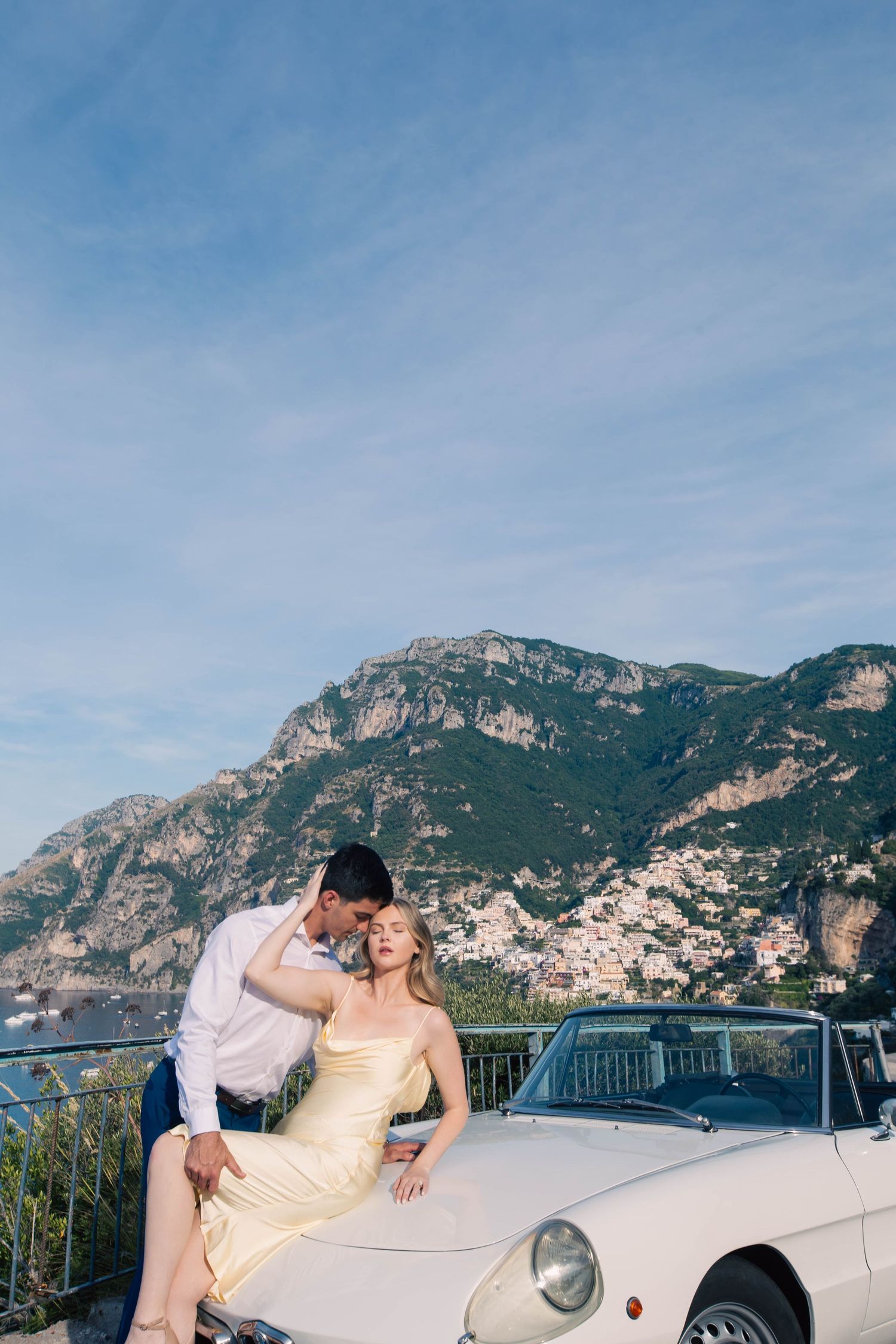 Couple posing with a vintage white convertible on the Amalfi Coast, with Positano village and mountains behind.