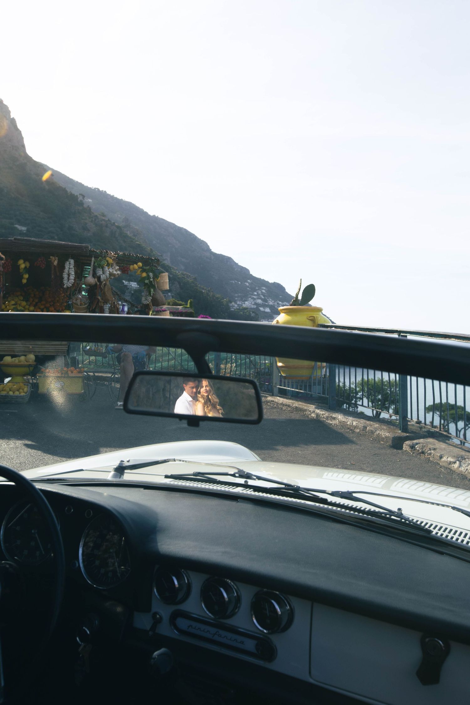 Smiling couple captured in vintage convertible car rearview mirror on sunny Amalfi Coast road.