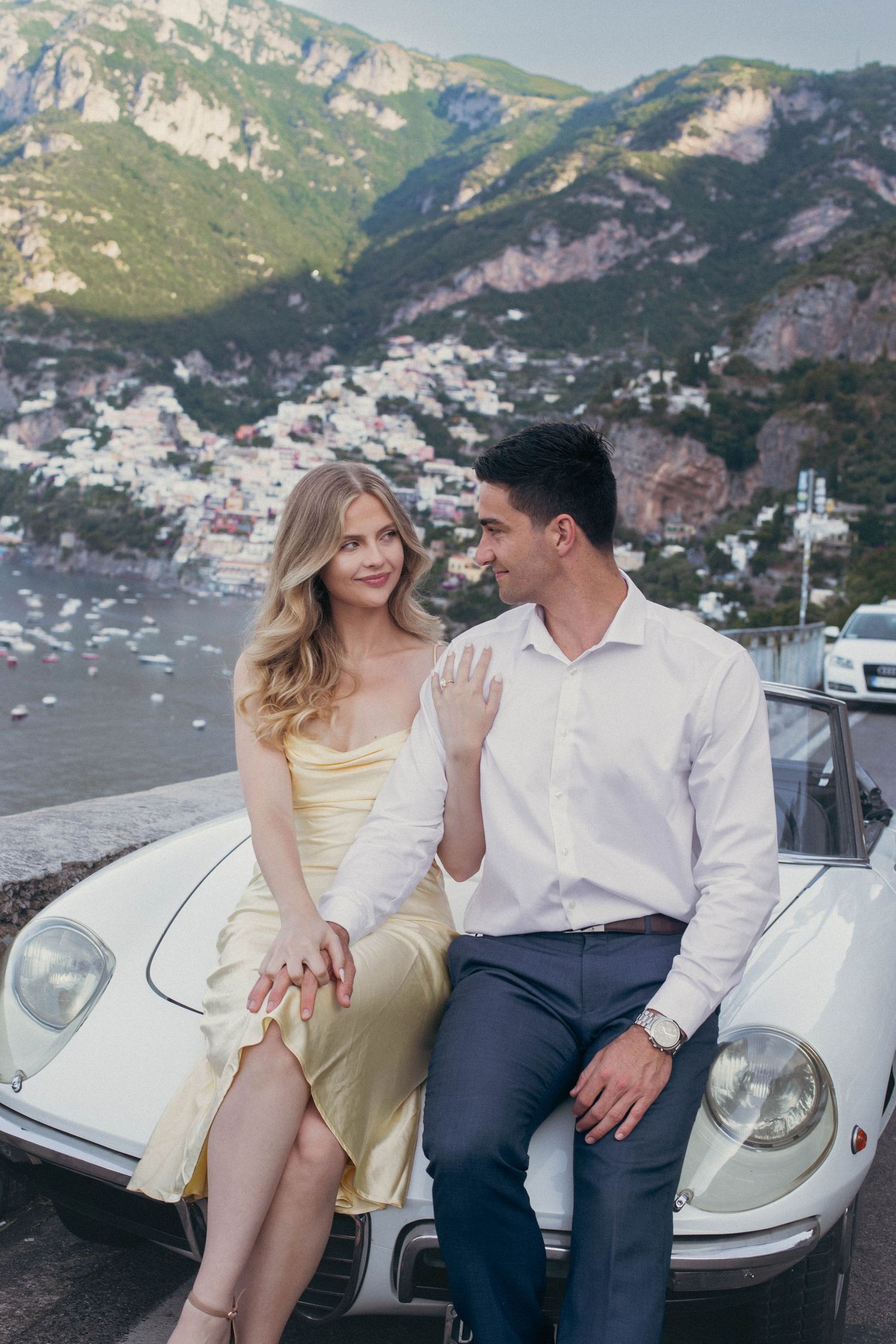 Elegant couple sitting on white classic Alfa Romeo convertible with scenic Positano coastline in background.