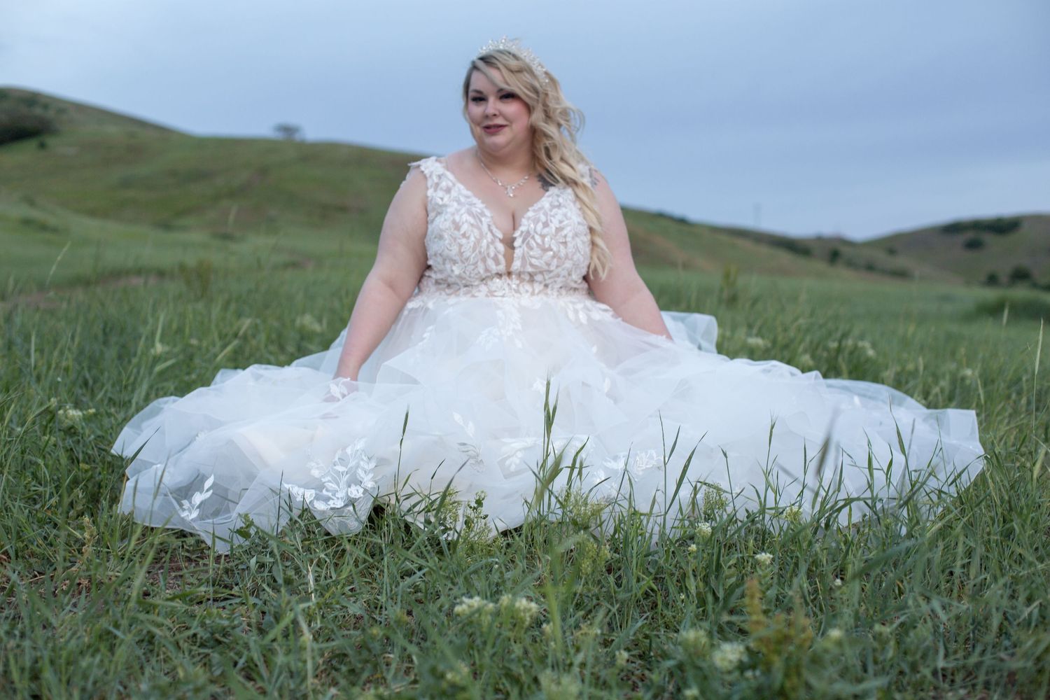 A white wedding dress with lace bodice spreads across green hillside grass at sunset.