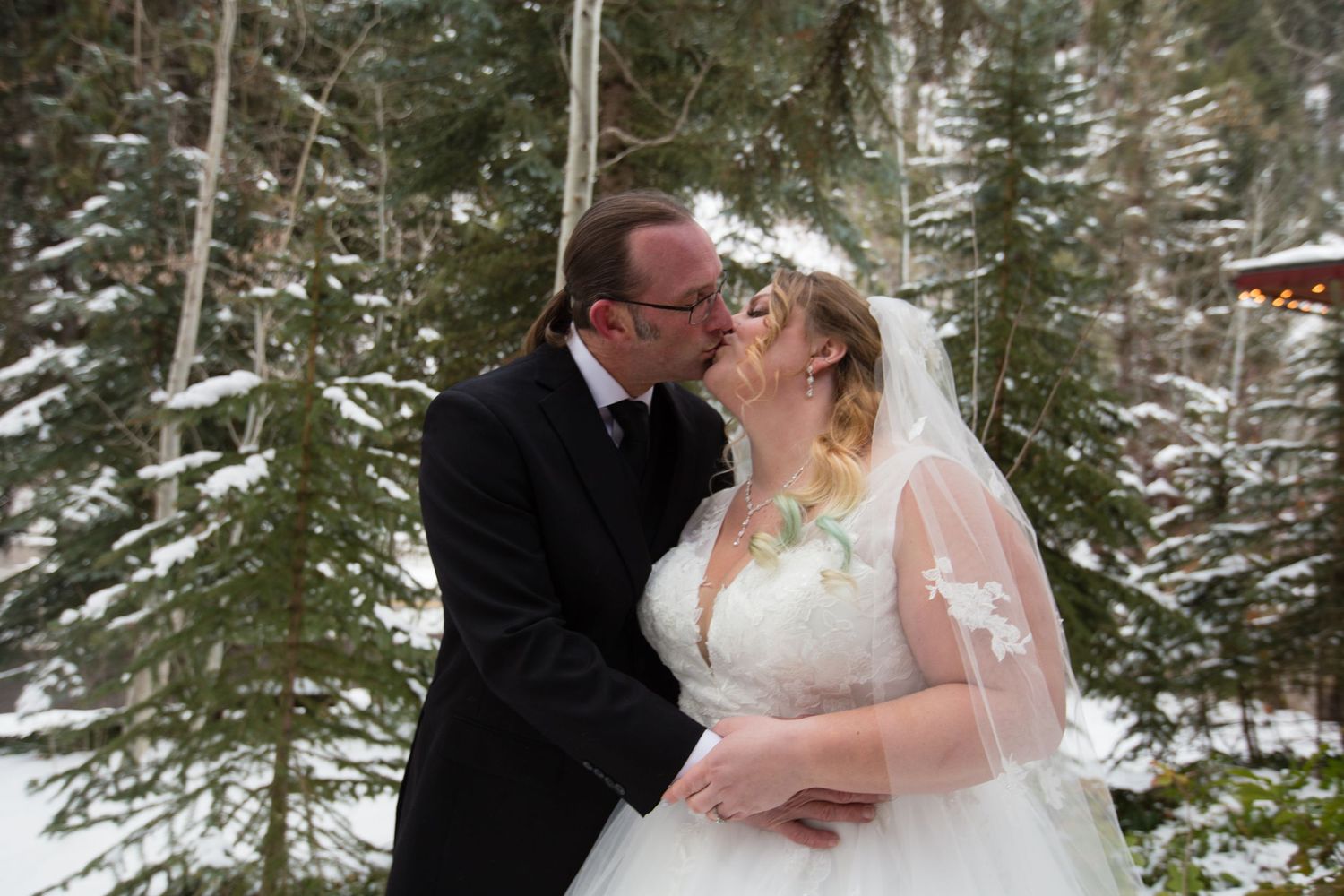 A tender embrace captured during a snowy winter wedding photoshoot among evergreen trees.
