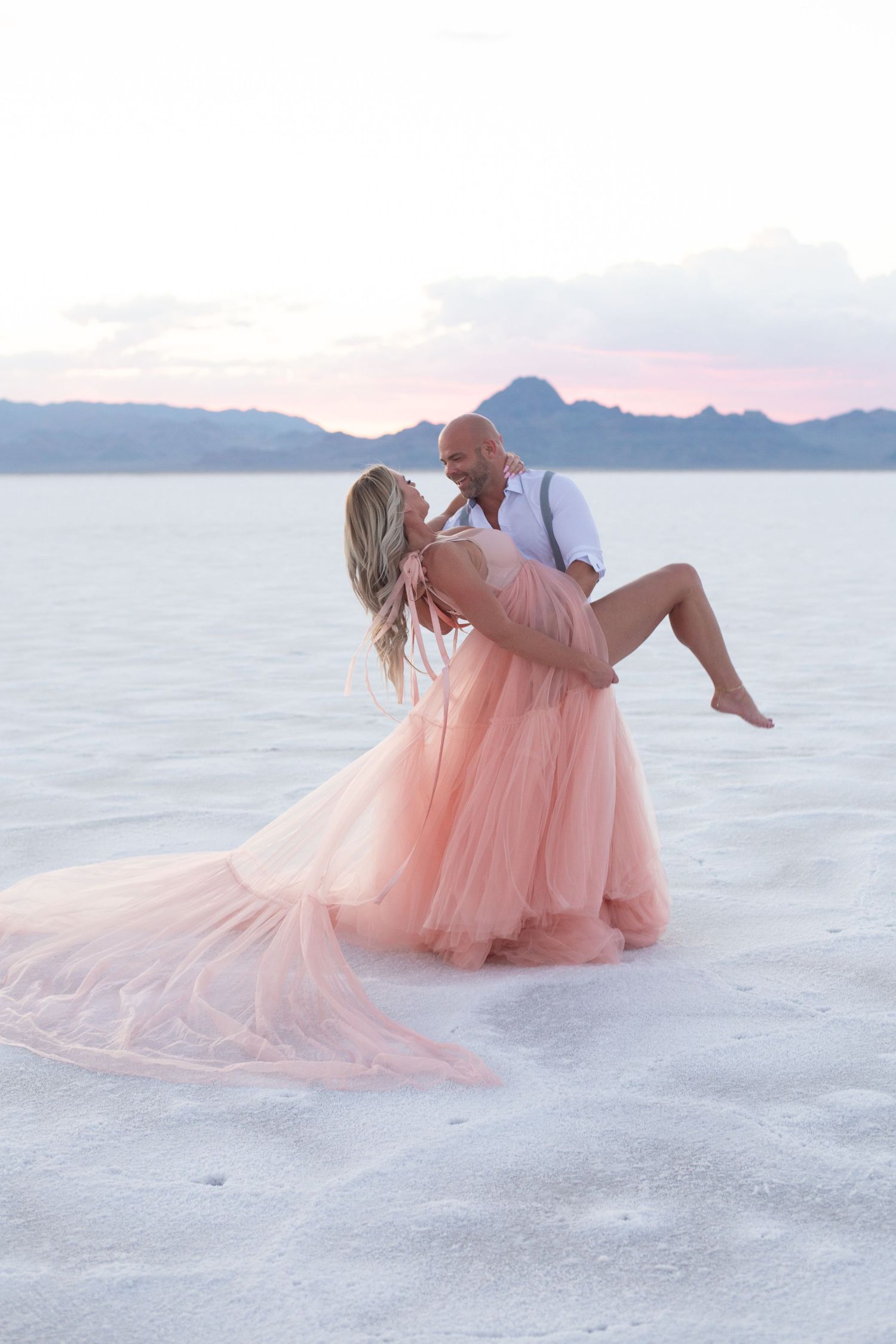 Romantic dip pose on salt flats with flowing pink dress against sunset mountains.