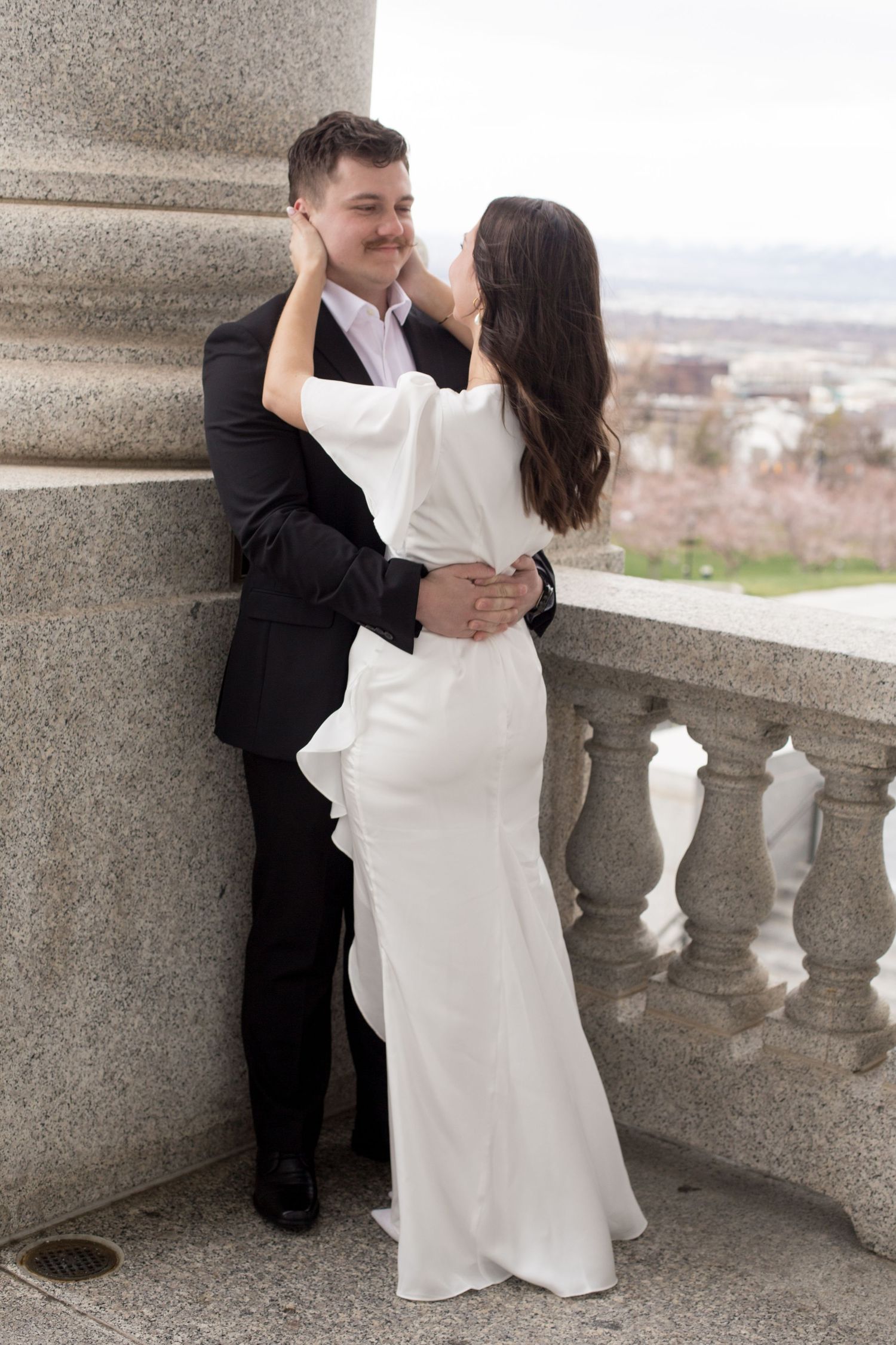 Bride in white gown embraces groom in black suit against stone balustrade with scenic backdrop.