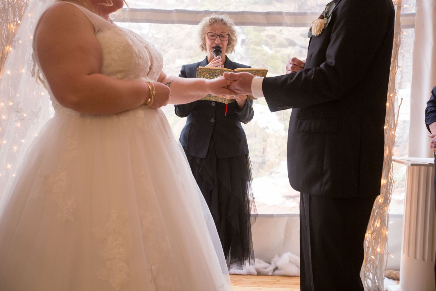 Bride and groom exchanging rings during indoor wedding ceremony with officiant and twinkling lights in background.