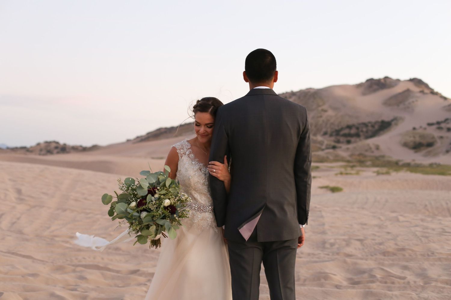 Couple in wedding attire standing on desert dunes at dusk.