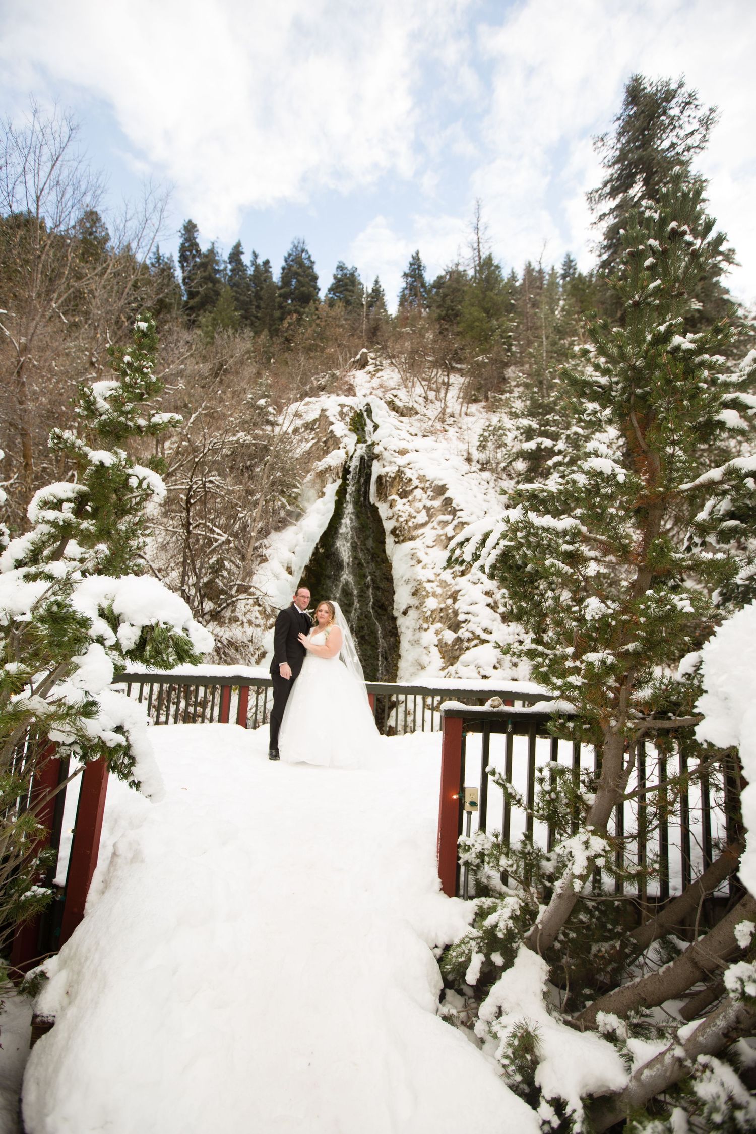 Wedding couple on snow-covered bridge with dramatic waterfall and evergreens in background.