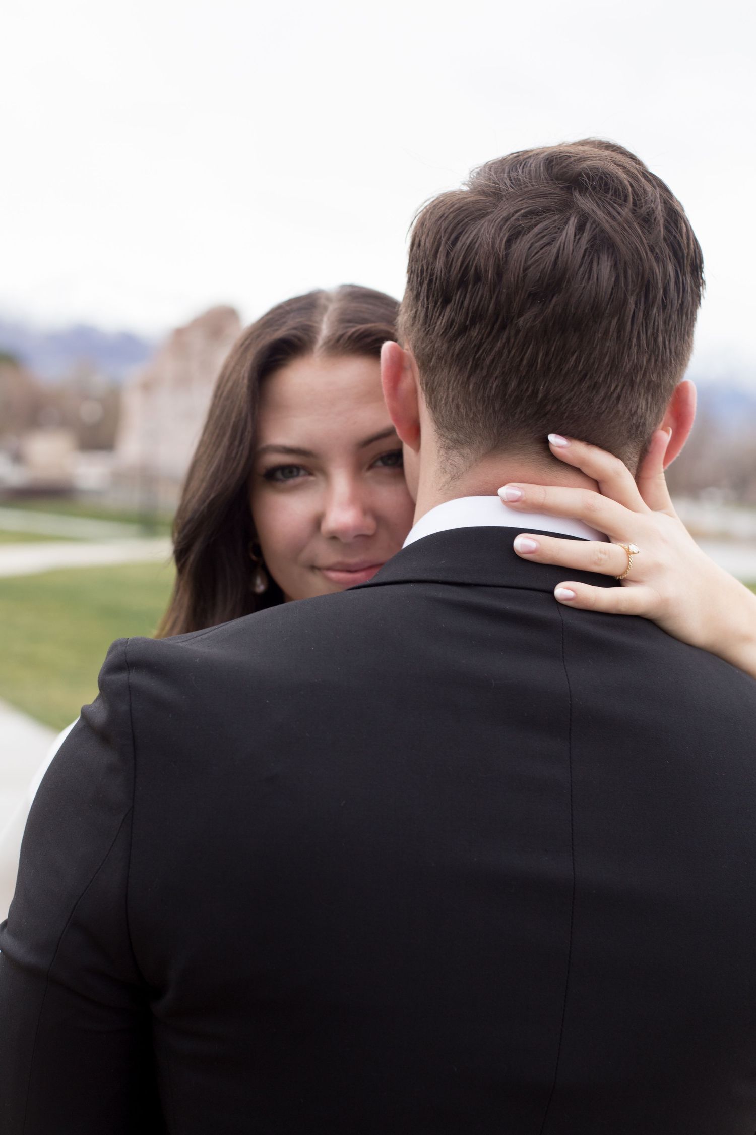 Bride embracing groom in black suit, smiling over his shoulder outdoors.