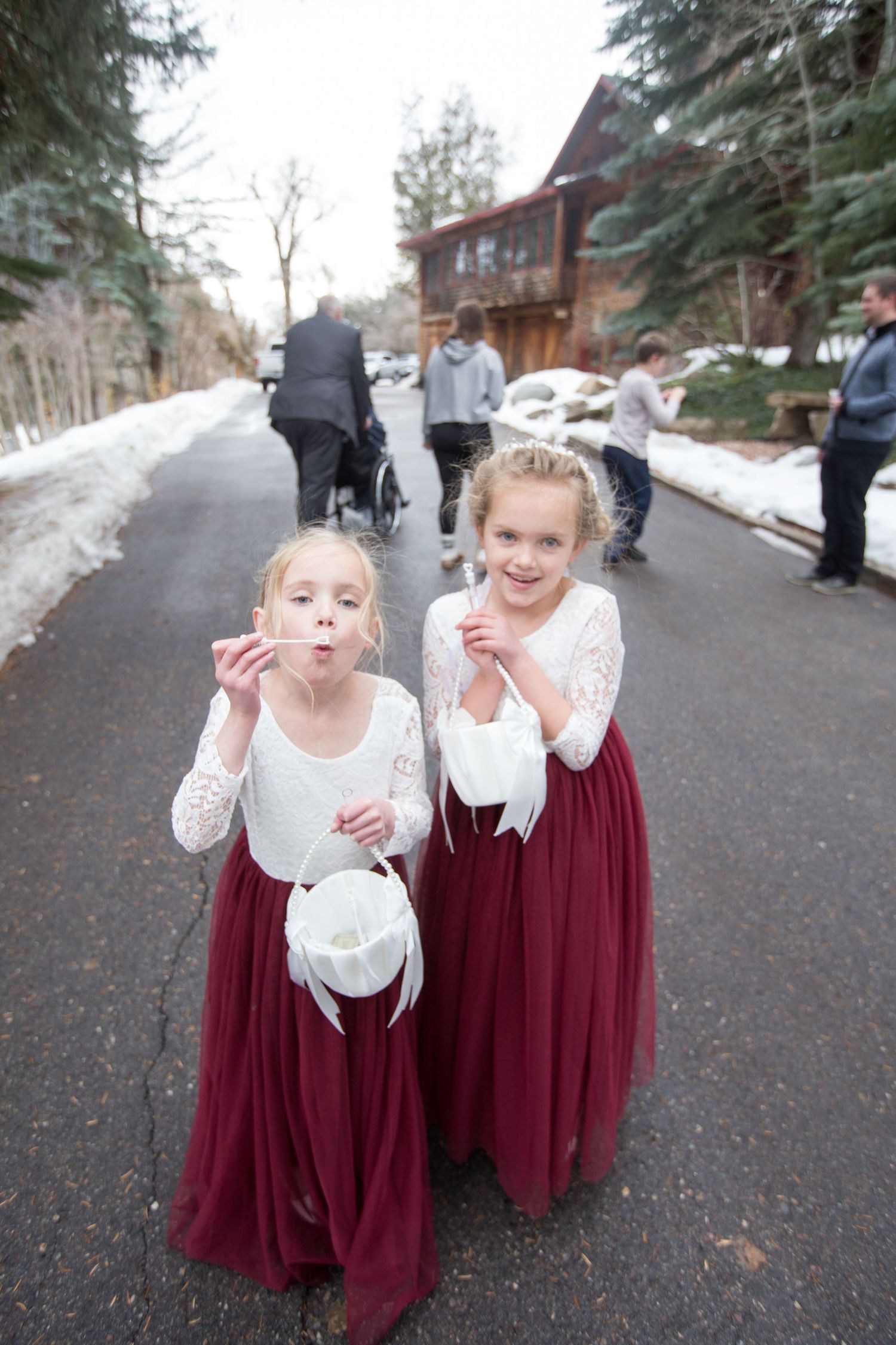 Two flower girls in white lace tops and burgundy tulle skirts blowing bubbles at winter wedding.