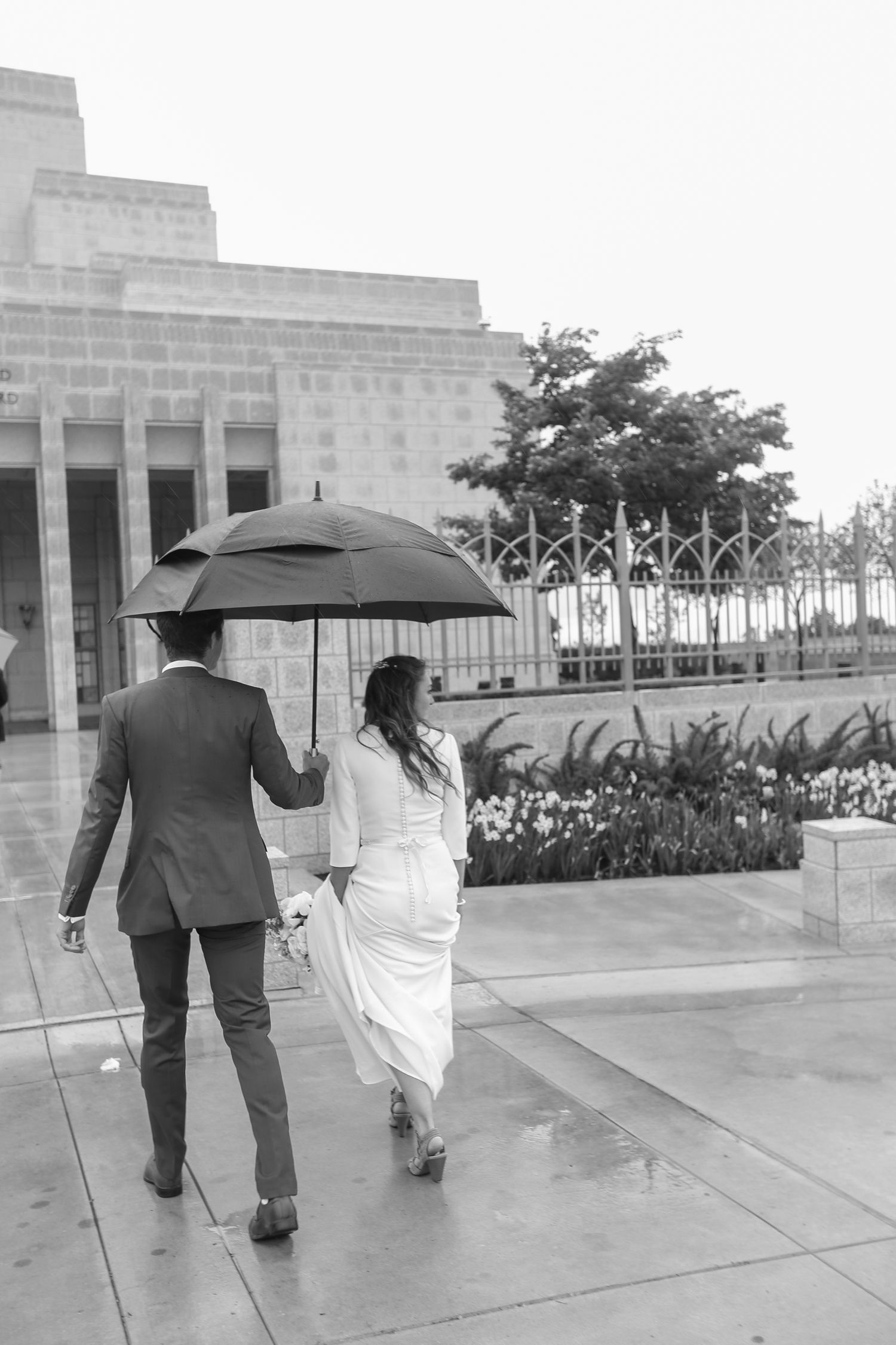 A couple walks with black umbrellas on a rainy day near a stone building.