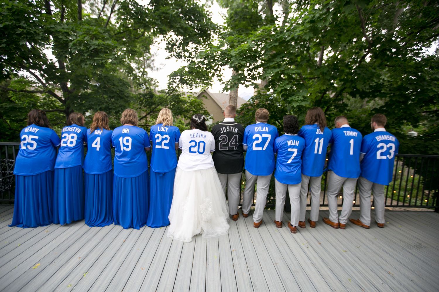 Wedding party lined up showing numbered jerseys in blue with bride in white dress on outdoor deck.