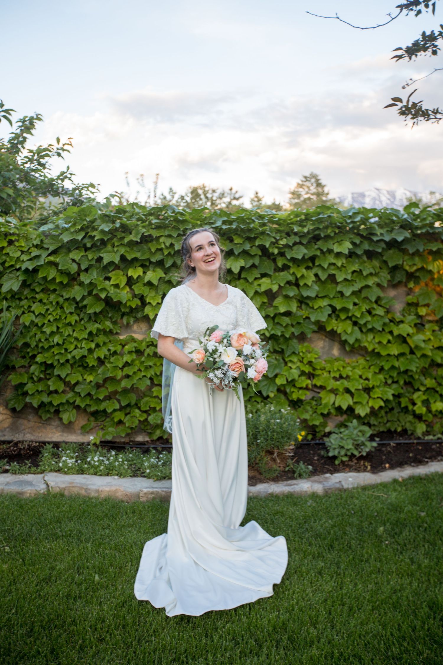 A bride in a flowing white dress poses on grass against an ivy-covered wall.