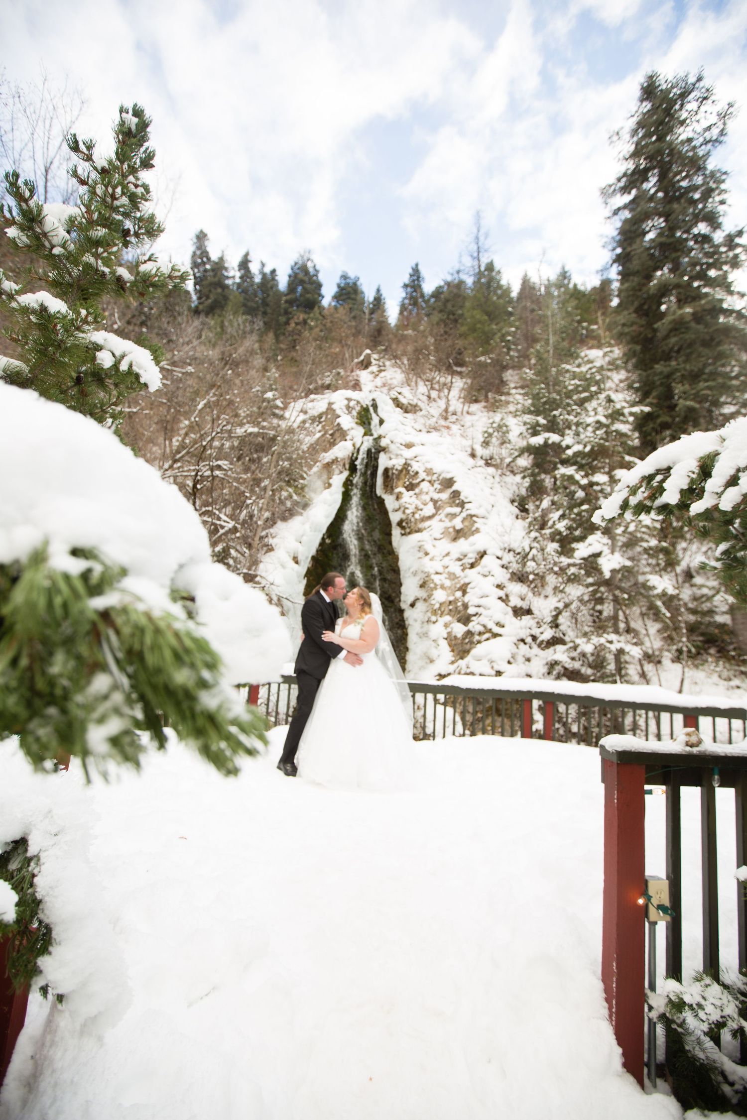 Newlyweds share an intimate moment on a wooden deck surrounded by snow-covered evergreens.