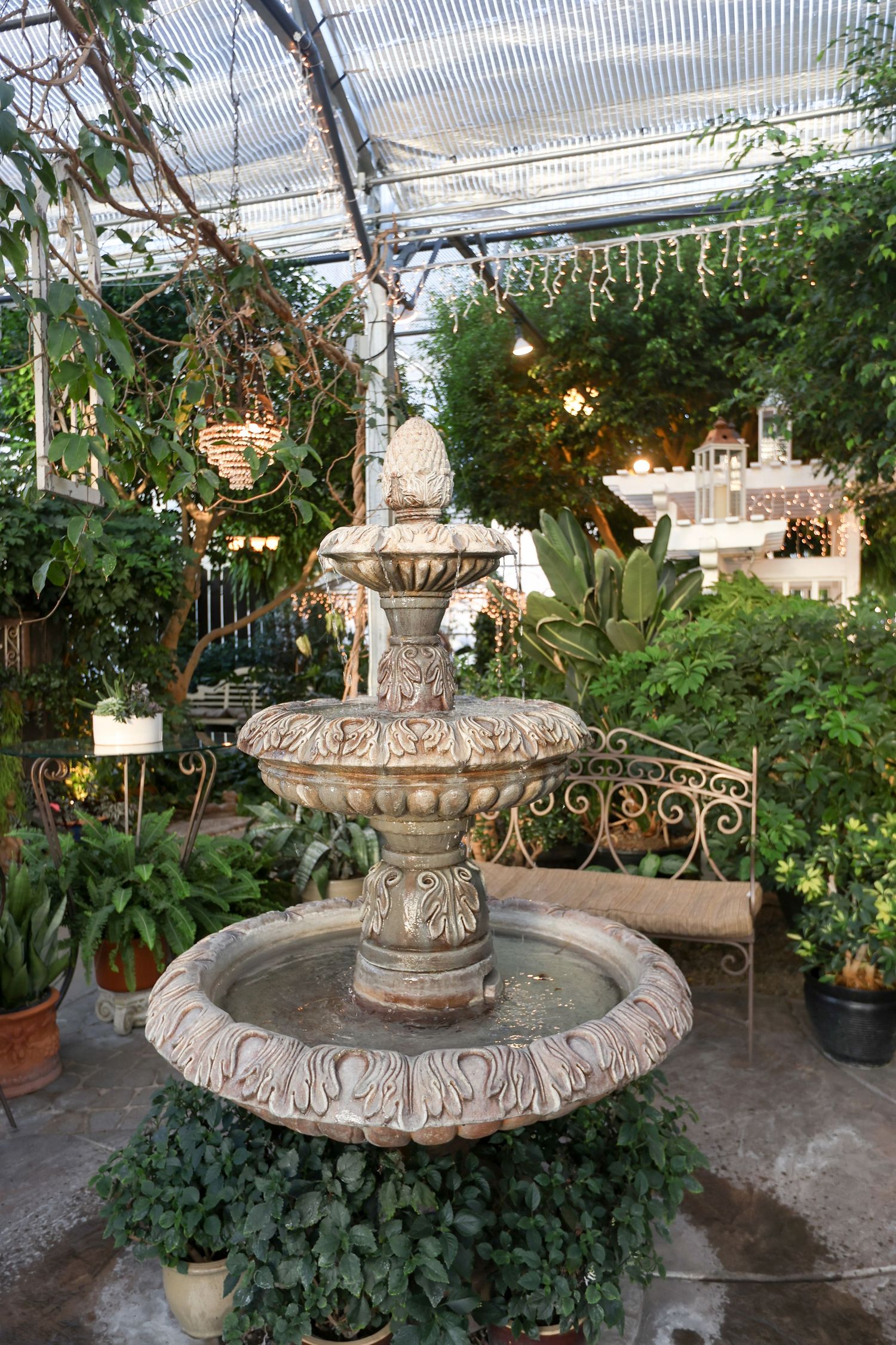 Ornate stone fountain surrounded by potted plants in a greenhouse setting.