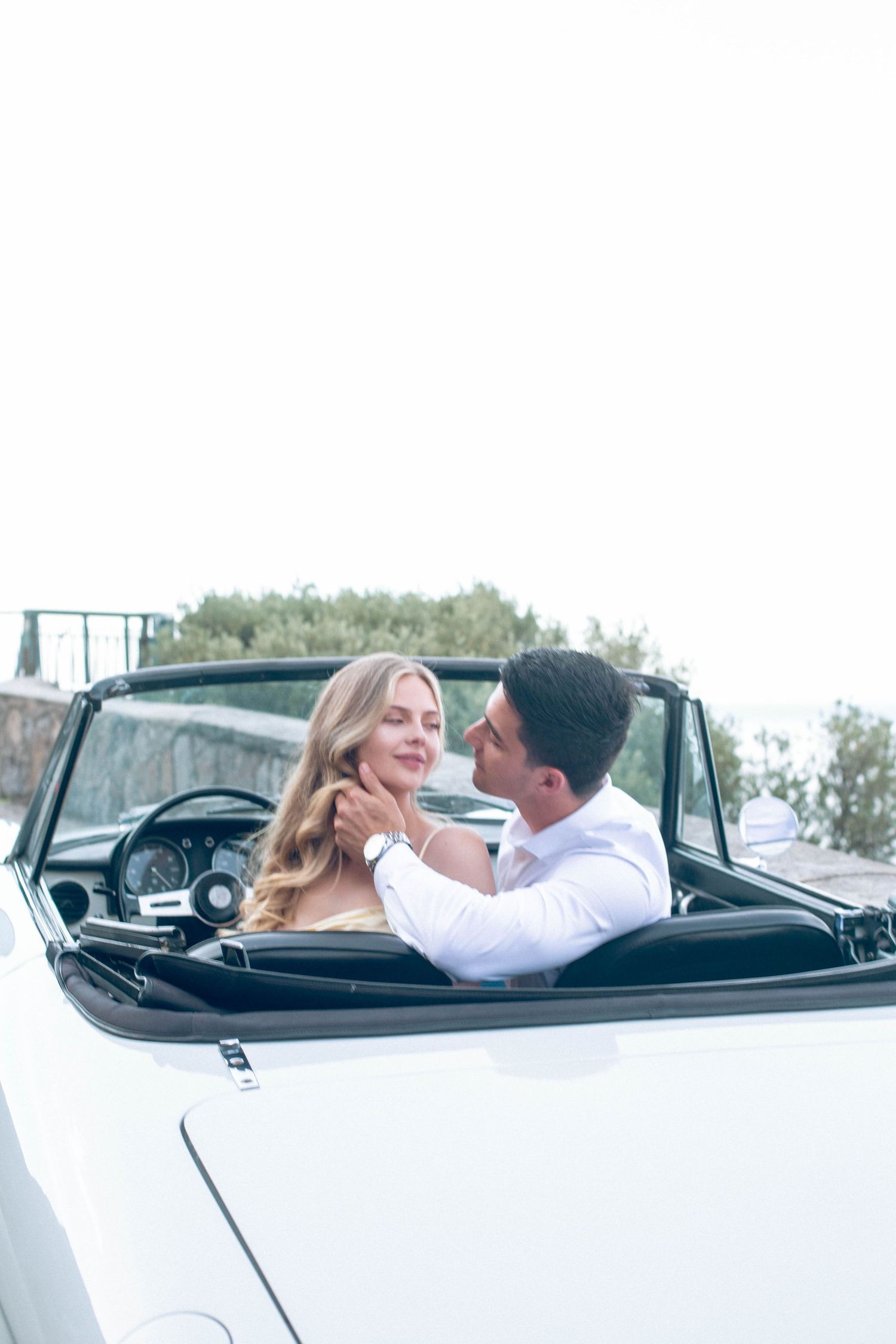 Romantic couple in a classic white convertible car, man tenderly holding womans face on the Amalfi Coast.