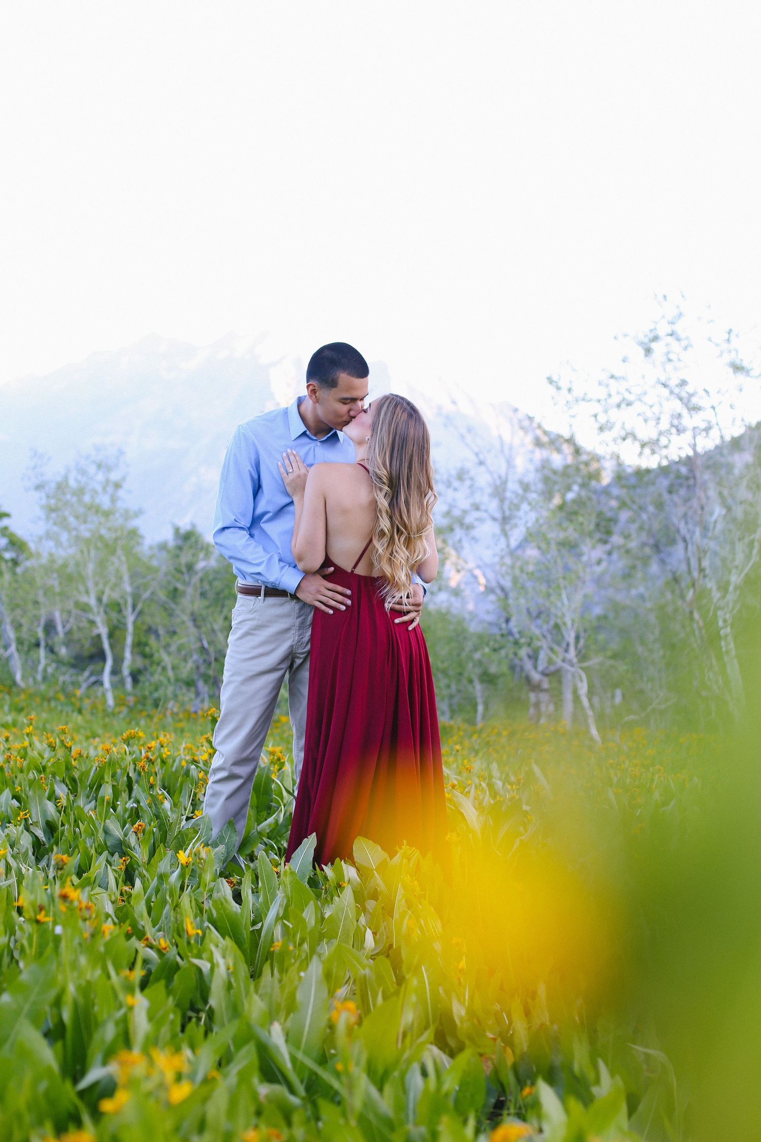Couple sharing a tender kiss in a vibrant wildflower field with mountain scenery in the background.