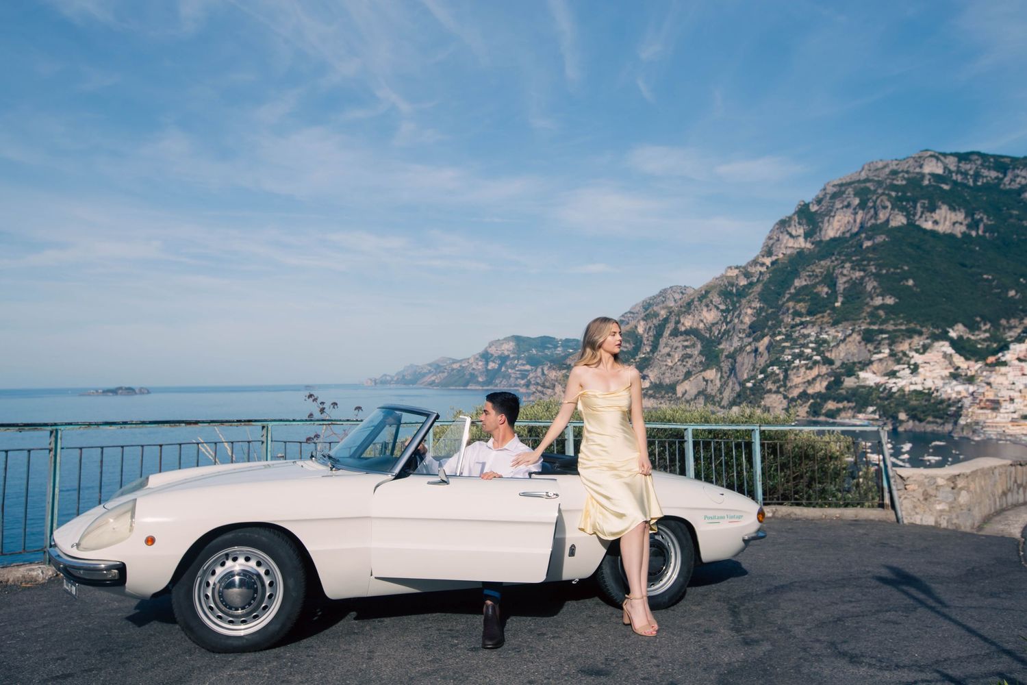Couple posing elegantly with white classic convertible on Amalfi Coast road with Positano village behind.