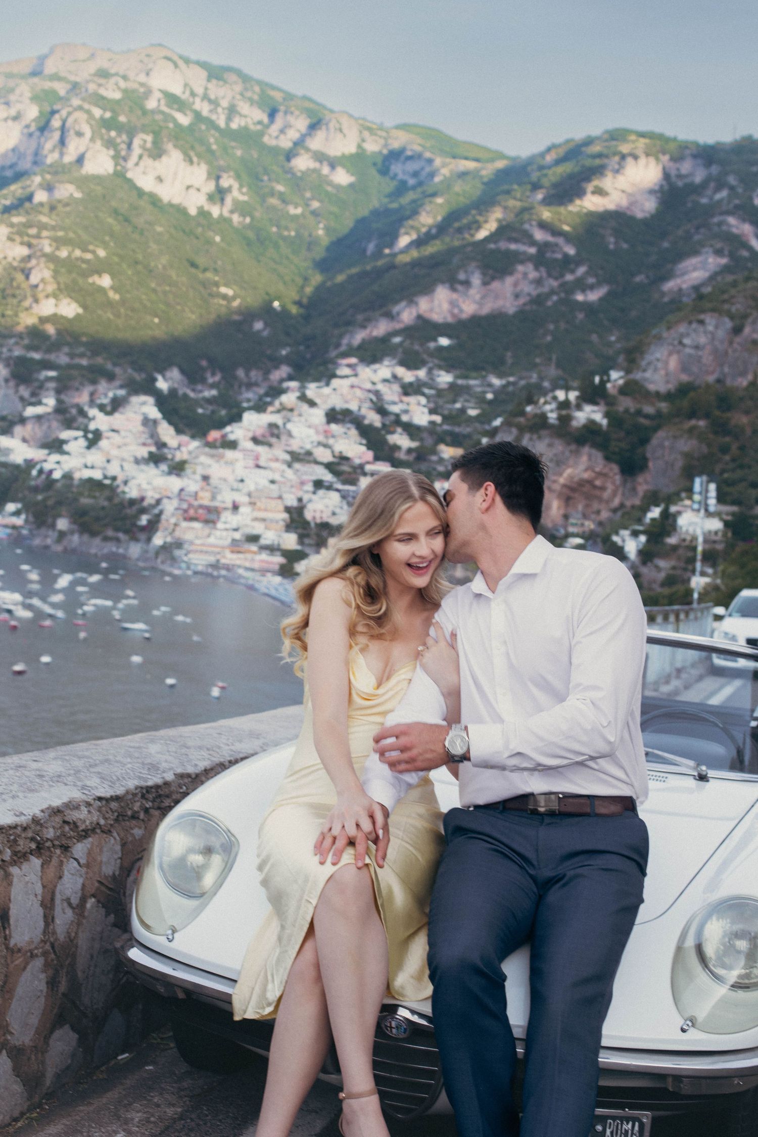 Couple leaning on vintage white car in Positano, Italy, with colorful cliffside village and Mediterranean sea backdrop.