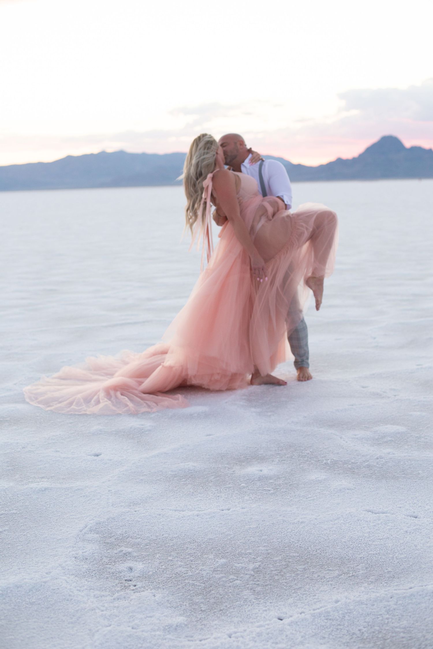 A couple poses romantically on white sand with mountains and water in the background at sunset.