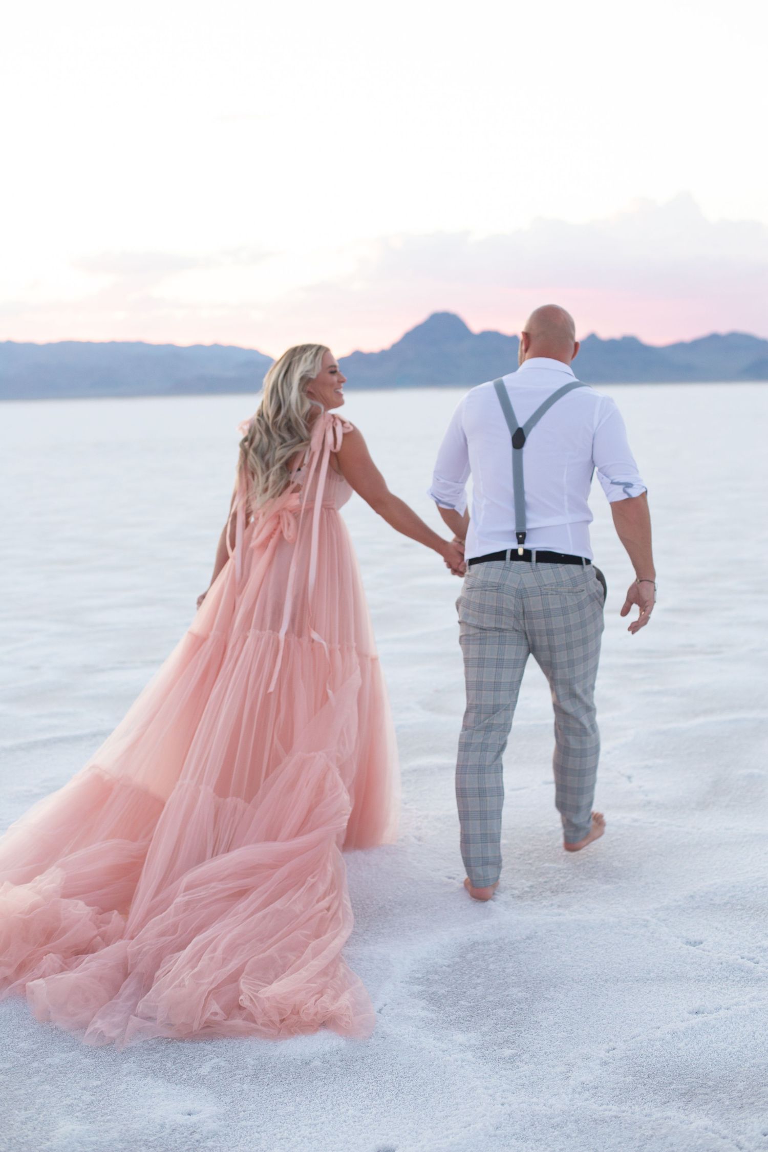 Couple walks barefoot on salt flats holding hands with pink dress trailing behind.