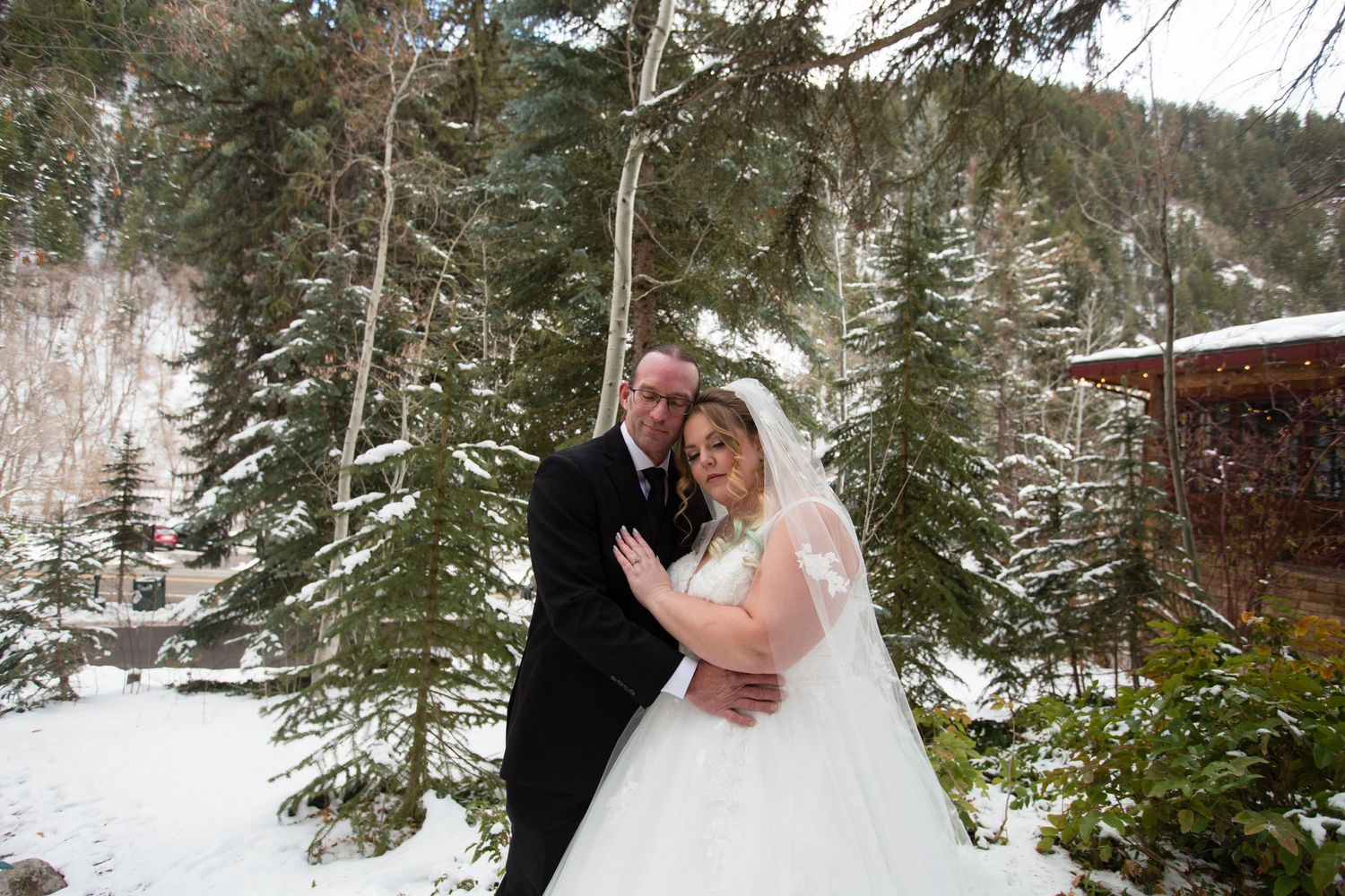 A romantic winter wedding portrait set against a backdrop of snow-covered pine trees and mountains.