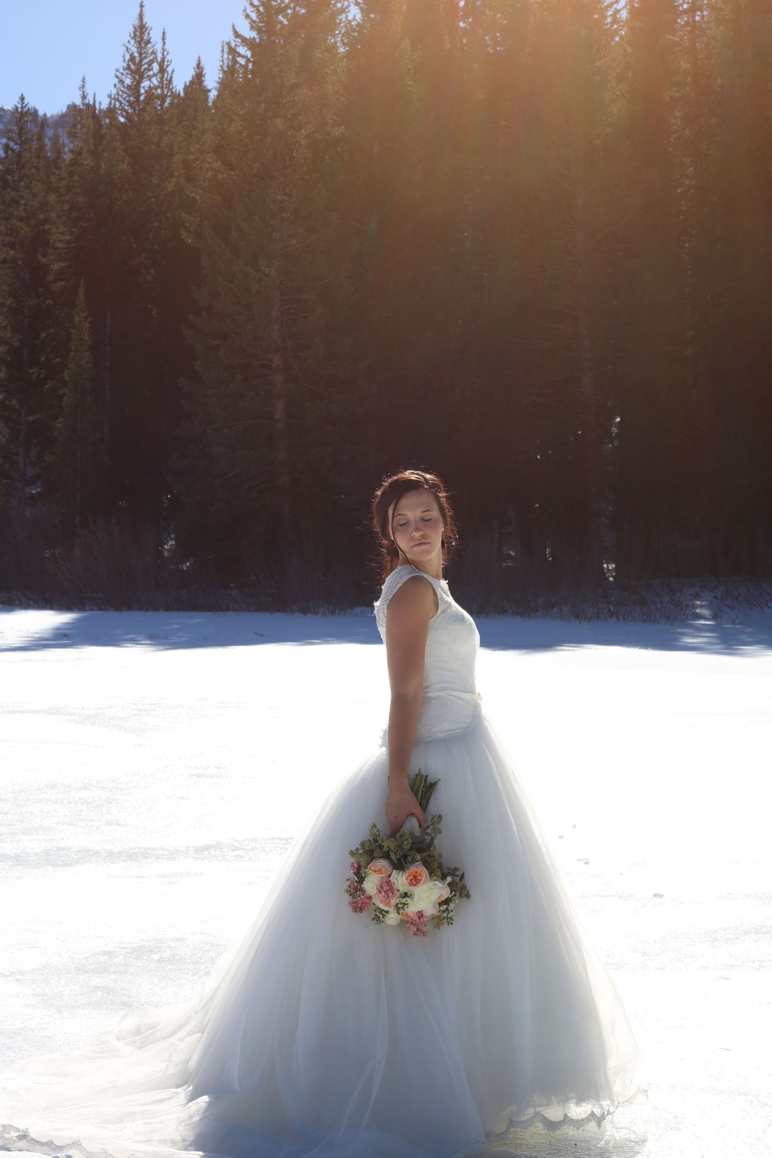Sunlit winter bridal portrait with bride holding bouquet in snowy forest setting.