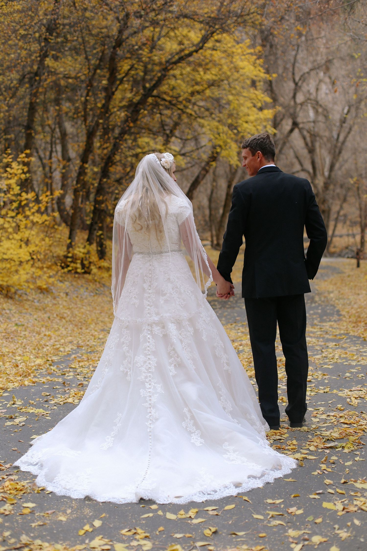 Wedding couple walking through autumn leaves with bride's long train and veil trailing behind.