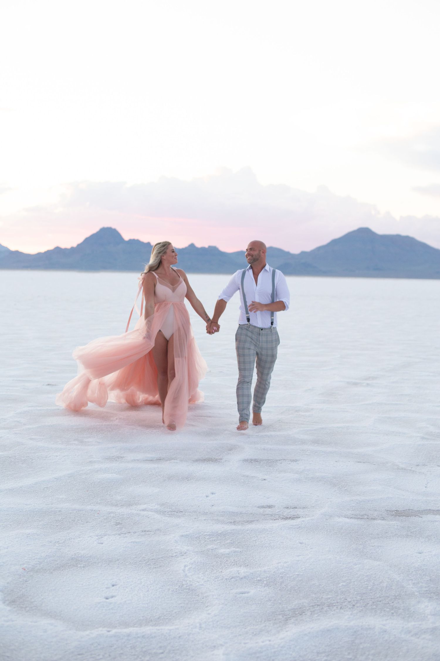 A couple walks hand in hand on white sand with mountains in background, bride wearing flowing pink gown.