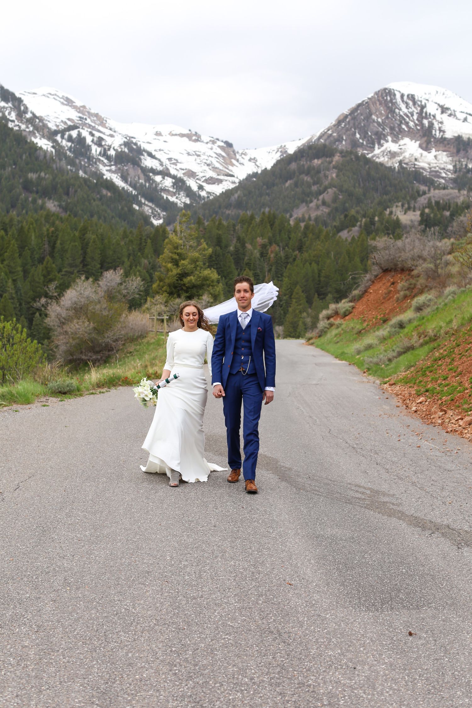 Newlyweds walk together on mountain road with snow-capped peaks in background.