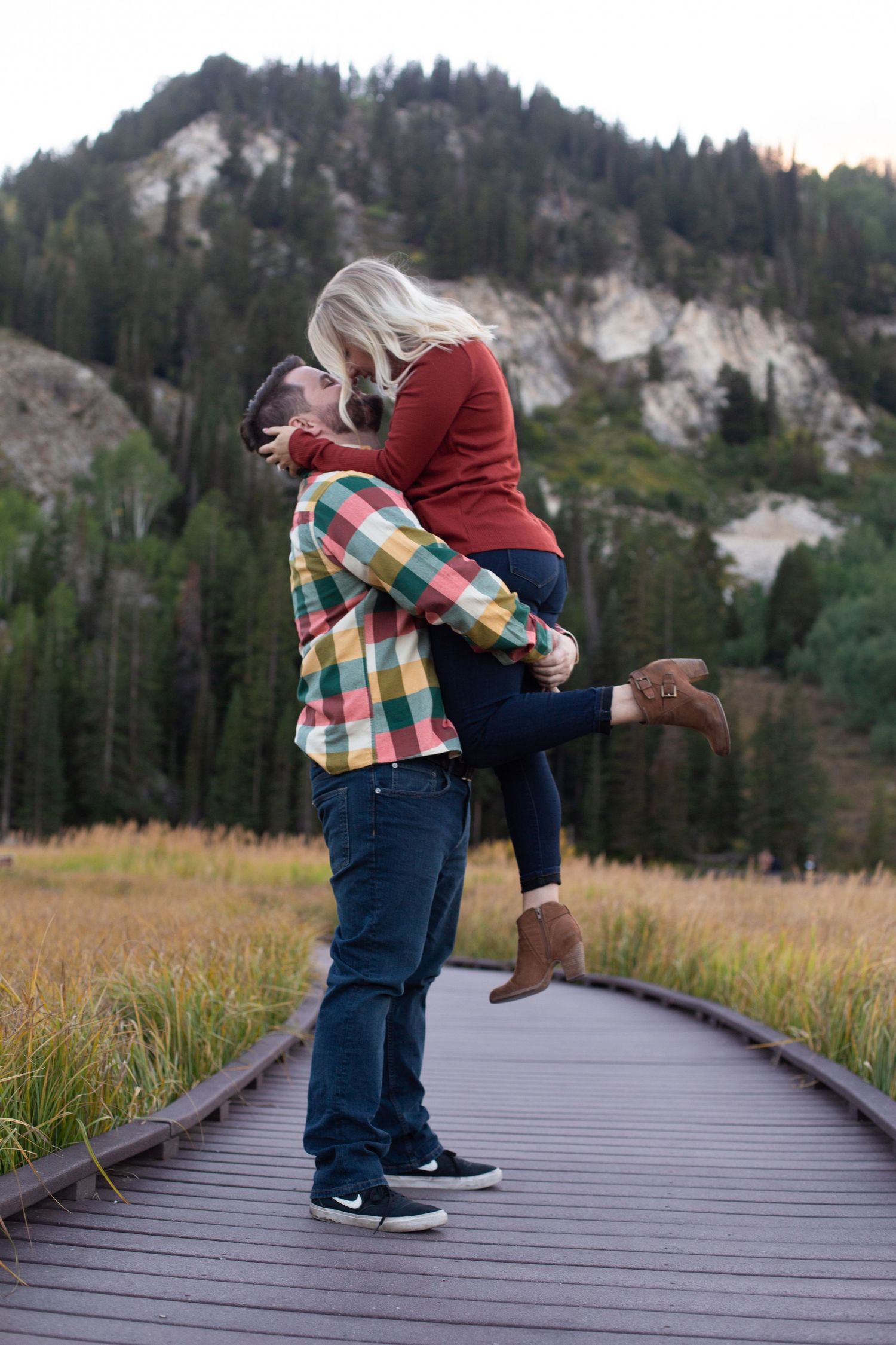 Couple in casual attire embracing on wooden boardwalk with mountain landscape behind.