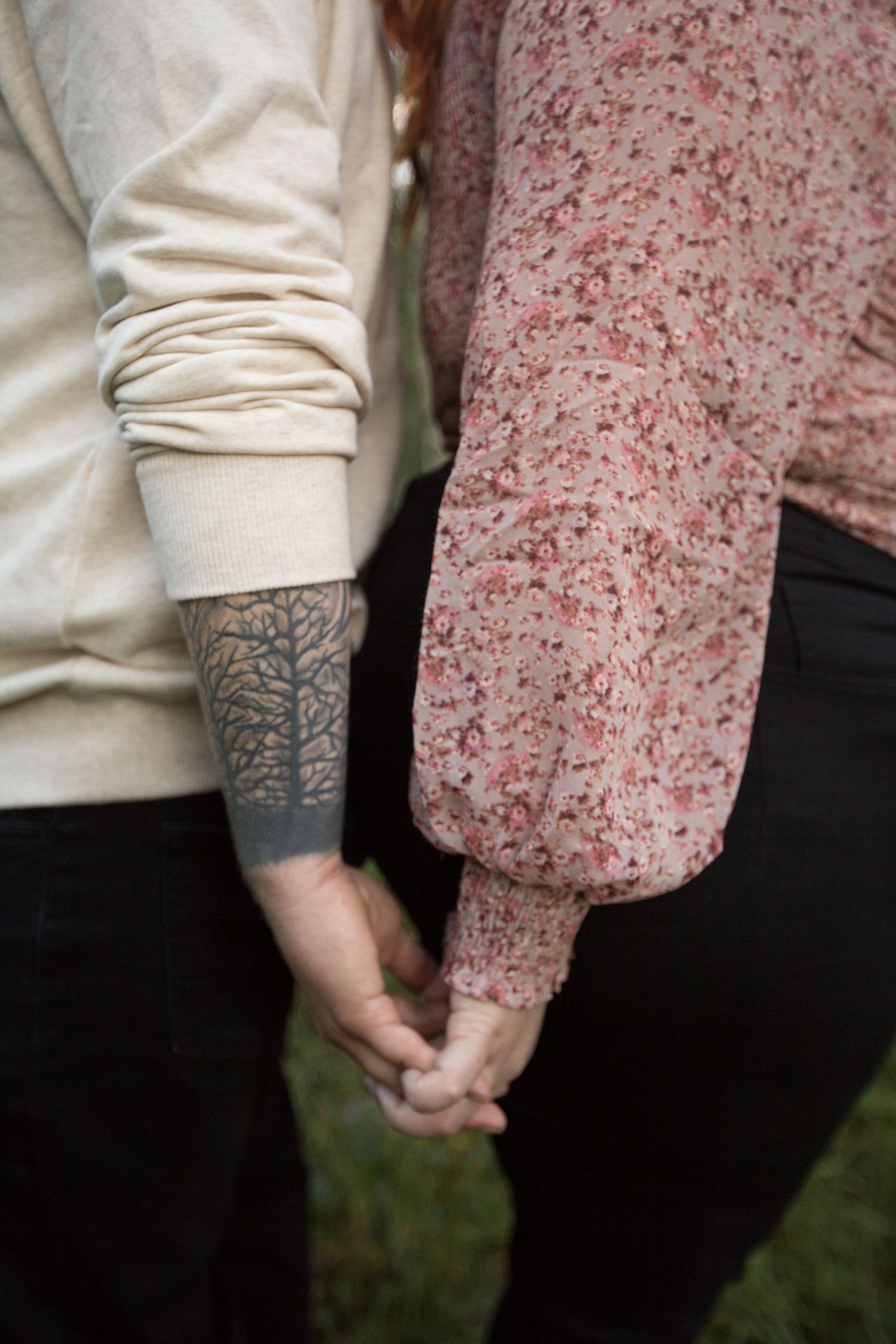 Close-up of two people holding hands, with one person wearing a beige sweater showing a detailed forearm tattoo.