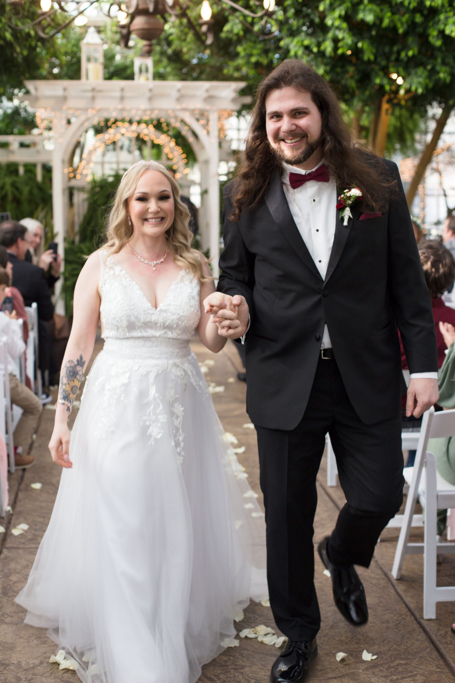 Newlyweds walking down aisle in greenhouse venue with white chairs and natural lighting.