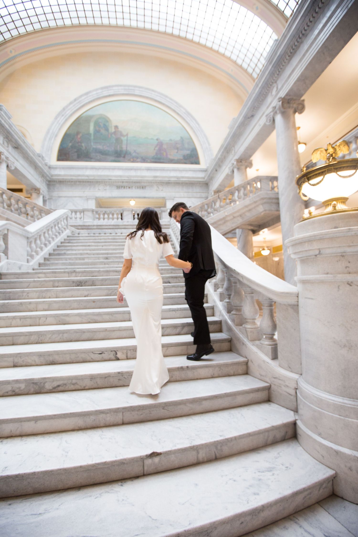 A couple ascending a grand marble staircase with ornate architectural details and vaulted ceiling.