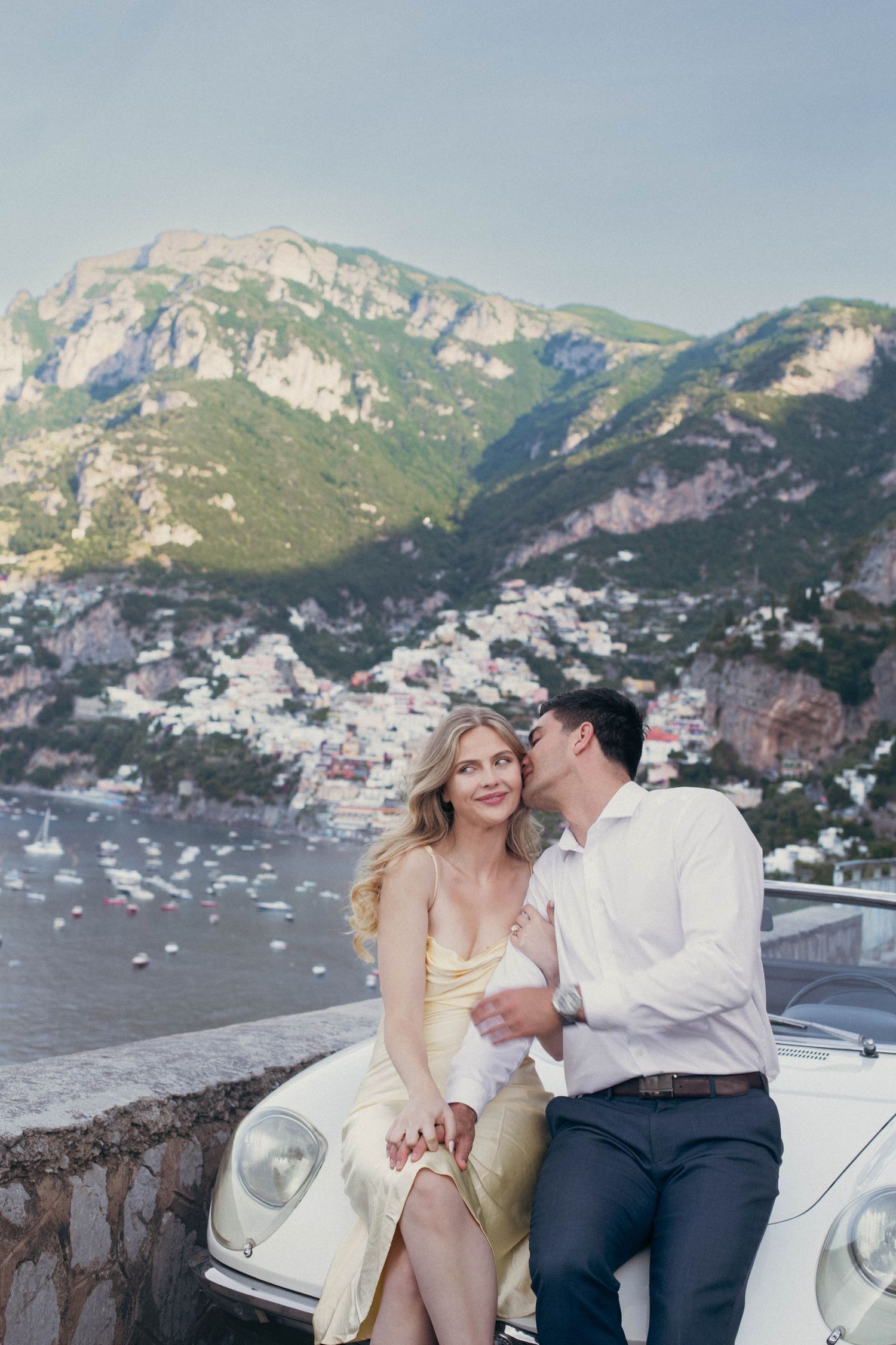 Elegant couple sharing intimate moment on classic white car overlooking scenic Positano coastal village Italy.