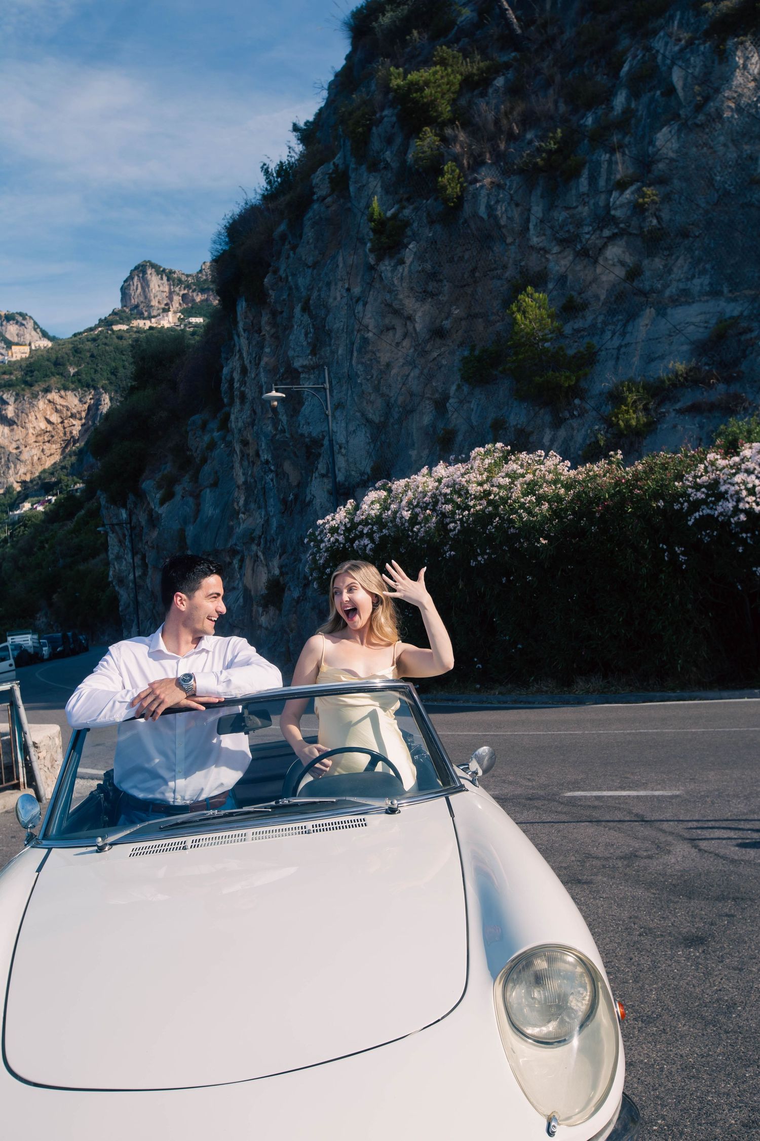 Excited couple riding in a classic white convertible along a scenic Amalfi Coast road with rocky cliffs.