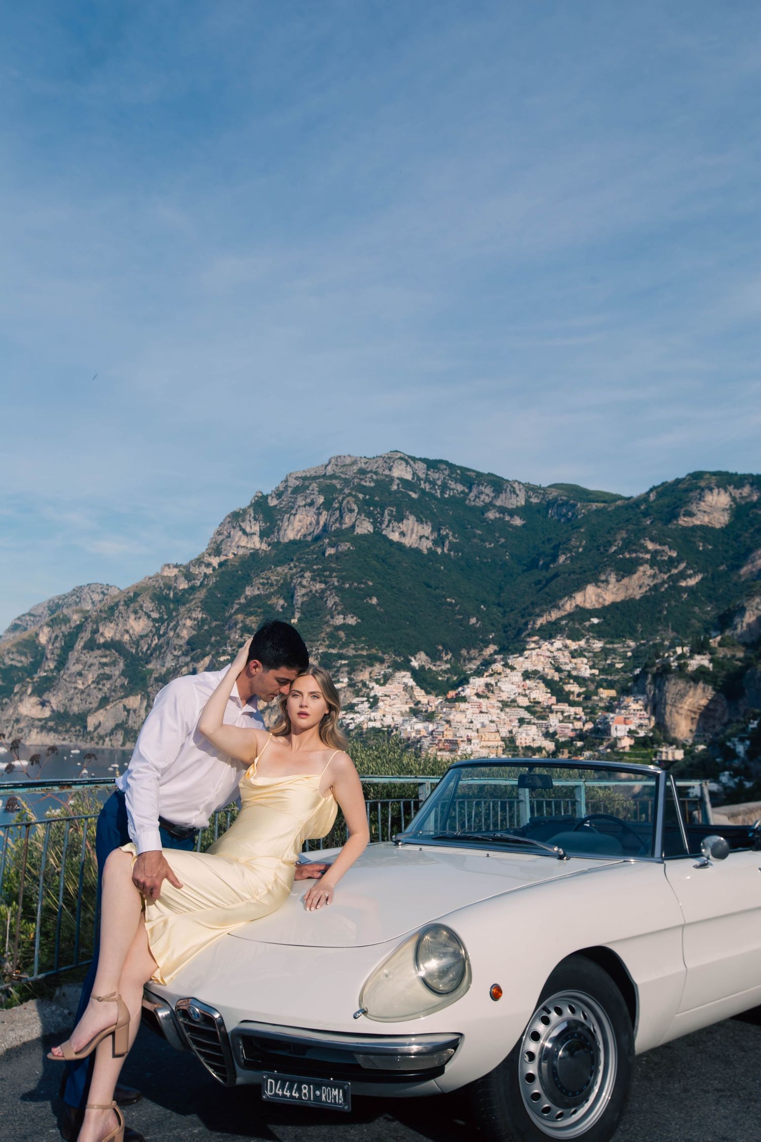 Couple embracing on hood of white Alfa Romeo convertible with Positano mountain village backdrop.
